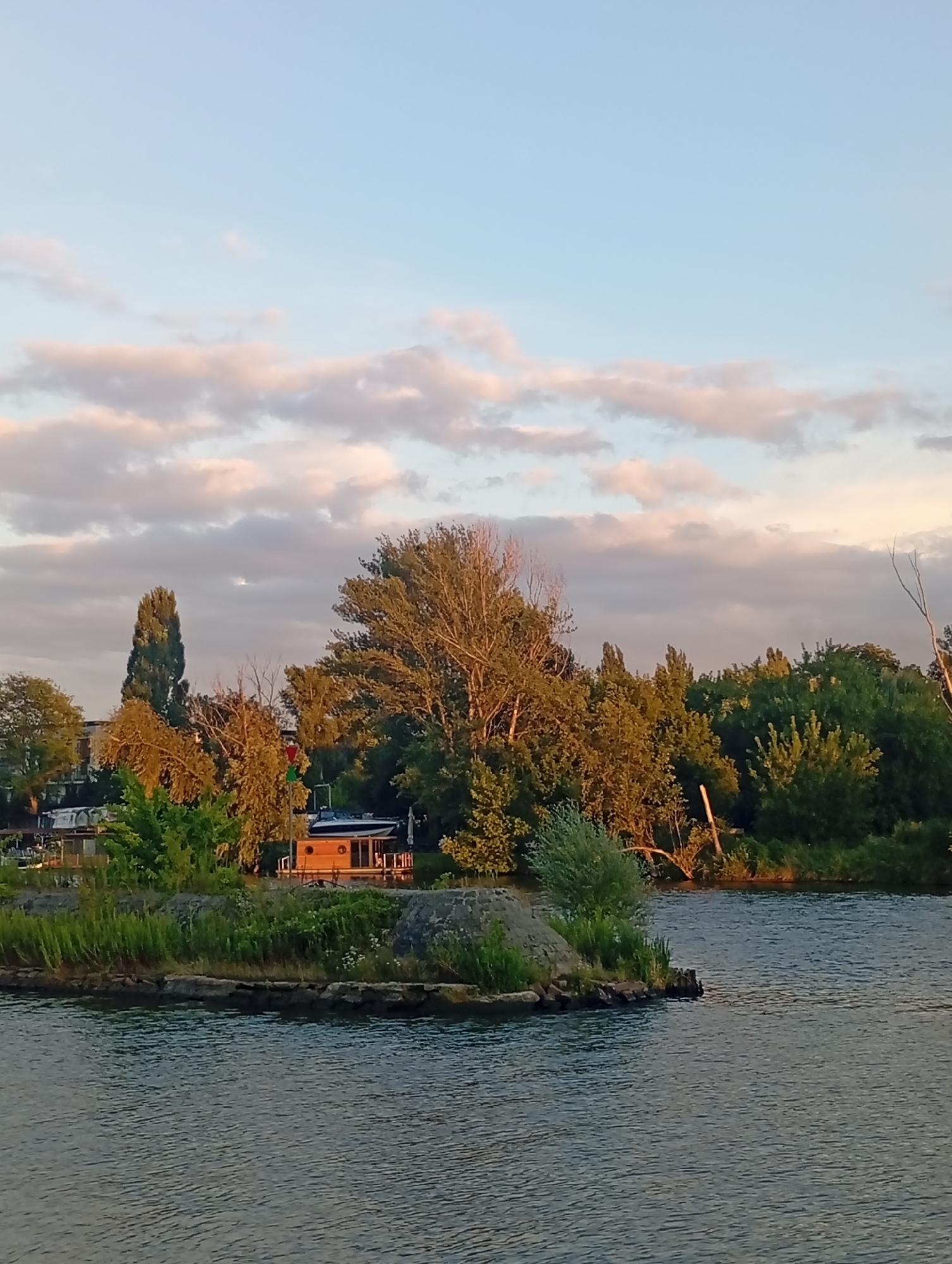 This image captures a serene waterside scene, likely taken from a boat or the bank of a river, overlooking a small island adorned with vegetation. The island is a focal point, featuring a charming wooden houseboat with circular windows, suggesting a peaceful, residential area. A navigational buoy with red and green lights stands near the houseboat, indicating a waterway. The background is a lush tapestry of trees and foliage, some of which are illuminated by the warm glow of sunset, painting the sky with soft hues of blue and pink. The water in the foreground reflects the ambient light, creating gentle ripples. Based on the presence of houseboats and the distinctive architecture visible in the distance, this scene is reminiscent of areas along the Odra River in Wrocław, Poland, possibly near the city center or a popular recreational spot. The image conveys a sense of tranquility and natural beauty within an urban landscape.