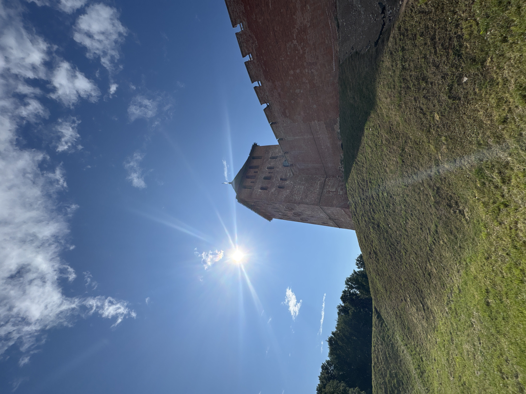 The image captures a view of the Veliky Novgorod Kremlin, a historic fortress in Veliky Novgorod, Russia. The foreground features a grassy, sloping embankment leading up towards the fortress walls. The walls themselves are constructed of weathered red brick, with crenellated battlements visible along the top. A tall, square tower, also made of red brick, rises from the wall, its windows appearing as dark rectangles. Above the Kremlin, the sky is a vibrant blue, dotted with scattered white clouds. The sun is shining brightly, creating prominent lens flares and rays of light that extend across the upper portion of the image. A line of dark green trees is visible along the lower left edge of the frame, beyond the embankment. The scene is bathed in bright sunlight, suggesting it was taken on a clear day. There are no people or animals visible in the image.