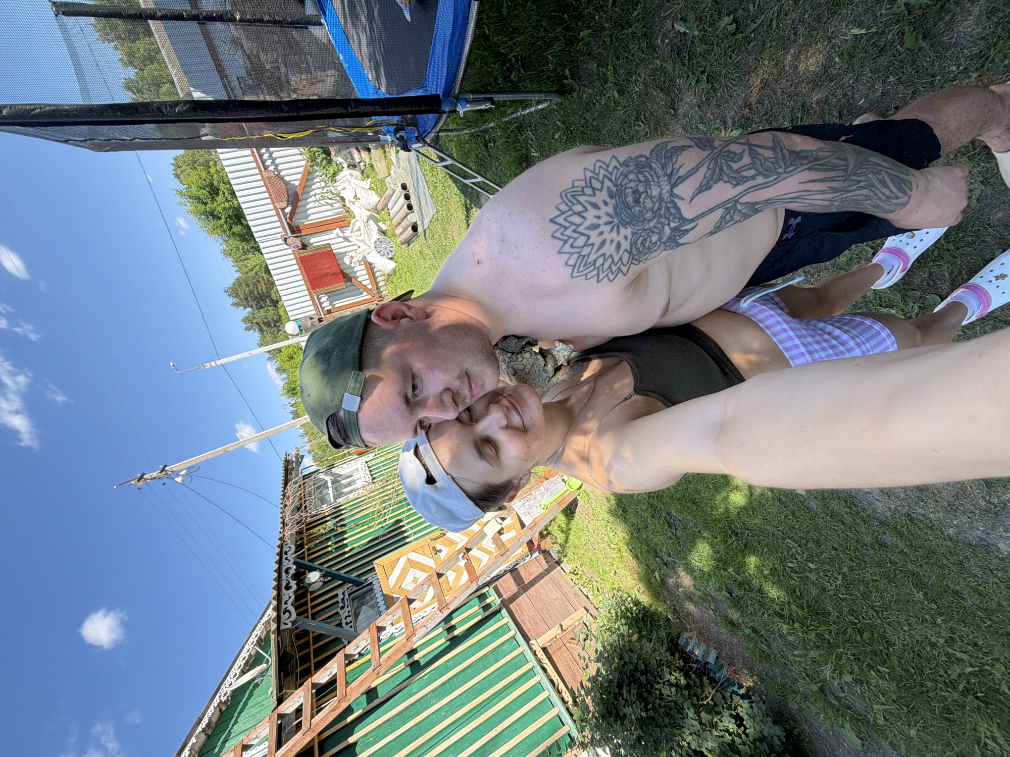 An outdoor, wide-angle, selfie photograph taken on a sunny day in the village of Krasny Mayak, Russia, features a man and a woman embracing. The man, shirtless and wearing a green baseball cap, has a prominent floral tattoo on his right arm. The woman, wearing a green tank top and a white baseball cap, is holding the camera. They are positioned in front of a rustic, green and yellow wooden house with a sloped roof and an external staircase. In the background, a trampoline with a black net is visible, along with a white metal fence, trees, and a utility pole with electrical wires. The ground is covered with green grass.