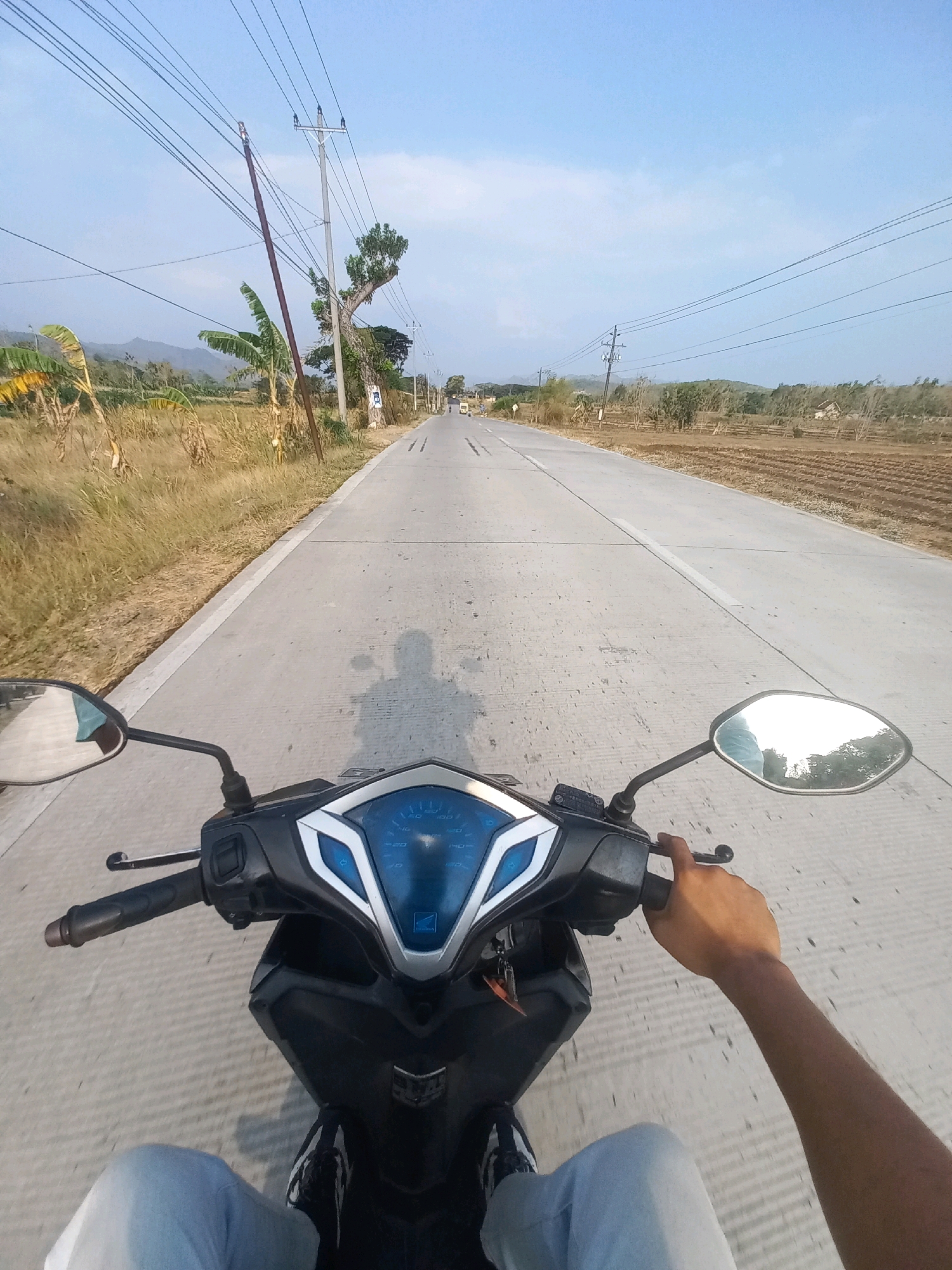 This is a first-person perspective shot of a person riding a motorcycle down a paved road in Baturetno, Indonesia. The road is grey and has white lines down the center. The rider's legs and feet, clad in light blue jeans and black and white sneakers, are visible in the foreground. Their right hand is on the motorcycle's handlebar, and their left arm is extended slightly to the side. The motorcycle's dashboard, with a blue display and a Honda logo, is also visible. The surrounding environment consists of dry grass and fields, with scattered trees and some banana plants on the left side of the road. Utility poles with numerous power lines stretch across the scene, and in the distance, hills are visible. The sky is clear blue with some light clouds. The overall lighting suggests it is daytime, likely late afternoon due to the long shadow of the rider cast on the road.