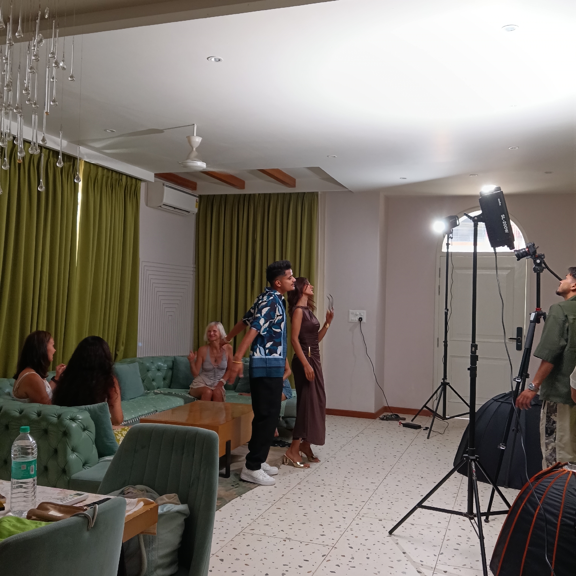 The scene is a room in Calangute, India, set up for what appears to be a video or photo shoot. Four women are seated on a teal-colored, tufted sofa with wooden coffee table in front of them, while a man in a blue patterned shirt and a woman in a brown dress stand nearby. The man is gesturing. A crew member stands near the door with professional lighting equipment. The floor is tiled with a speckled pattern. There are green curtains and a decorative light fixture hanging from the ceiling. Other furniture includes a teal dining chair and a table with items on it, including a water bottle.