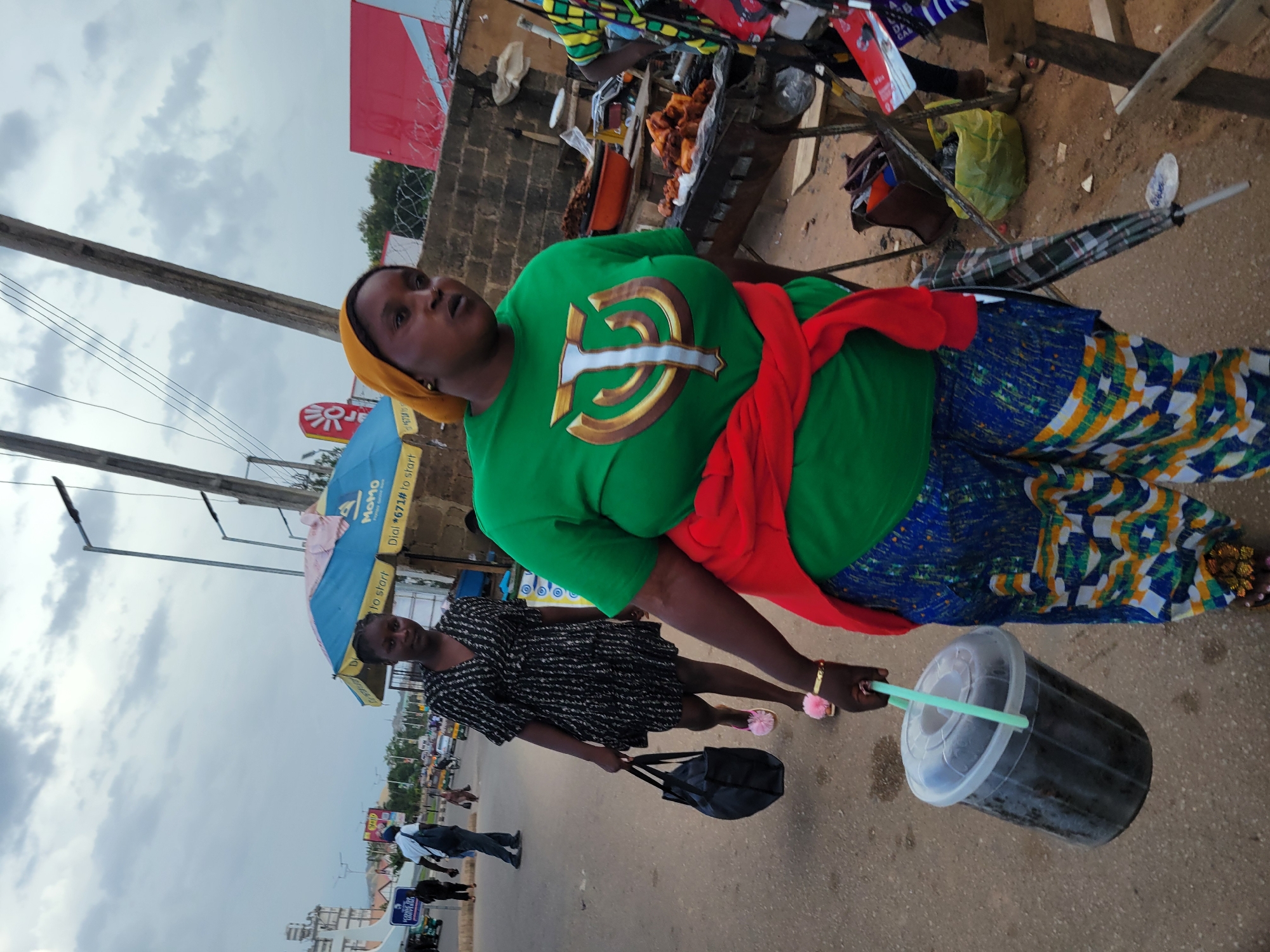 The image captures a street scene in Jos, Nigeria, with a focus on two women. The foreground is dominated by a woman wearing a green t-shirt with a gold emblem, a yellow headscarf, and a vibrant, patterned skirt. A red cloth is tied around her waist, and she is holding a large plastic cup with a straw. Her expression is directed upwards and slightly to the side, suggesting she might be looking at something or someone off-camera.
Behind her, another woman is visible, wearing a black and white patterned dress and pink fluffy slippers. She is carrying a black tote bag and appears to be walking. An umbrella, blue and yellow with some text on it, is partially visible in the background, possibly indicating a stall or vendor.
The setting appears to be an outdoor marketplace or busy street, with buildings and other people in the distance. The ground is paved, and there are visible power lines and streetlights overhead. The sky is overcast with clouds, suggesting a potentially gloomy or humid day. The overall impression is one of everyday life in a Nigerian urban environment.
There is some visible text on the blue and yellow umbrella, which includes "Momo" and "Dial #671*". Other signs and text are present in the background but are not clearly legible. The image seems to be taken during daylight hours, but the cloudy sky obscures direct sunlight. The activity taking place appears to be people going about their daily business, possibly shopping or commuting.