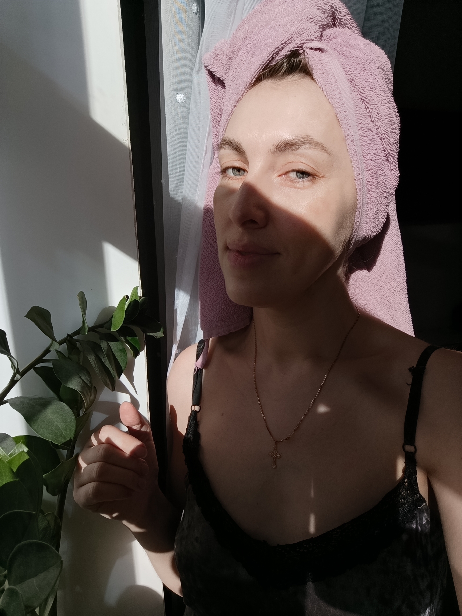 A woman with a pink towel wrapped around her head stands in a room next to a window. The window is on the left side of the image, with a white frame and sheer white curtains partially visible. A green leafy plant is positioned to the left of the woman. The woman wears a dark-colored camisole with lace trim and a gold necklace with a cross pendant. Sunlight is casting harsh shadows across her face and body. The location is unspecified in the image but the user asked about the city of Chaikovsky, Russia. The image does not include that information.