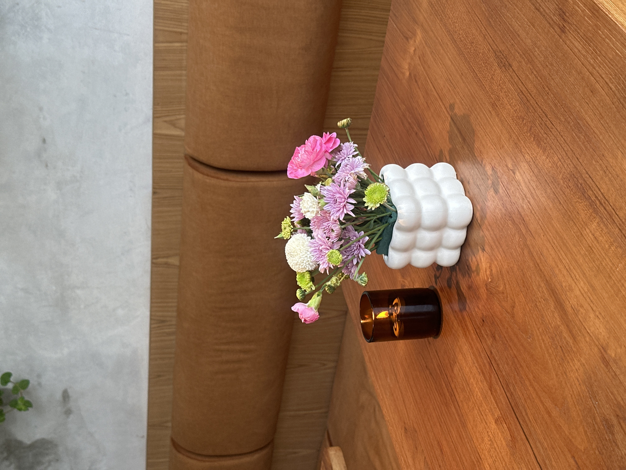 This image shows a close-up view of a wooden table in a cafe located in North Kuta, Indonesia. On the table, a lit candle in a dark glass holder sits to the left, and a white, bubble-shaped vase filled with a bouquet of pink, purple, and green flowers is placed to the right. In the background, a tufted brown leather sofa is visible, along with a textured grey wall. The overall ambiance is cozy and inviting, suggesting a relaxed setting within the urban environment of North Kuta.