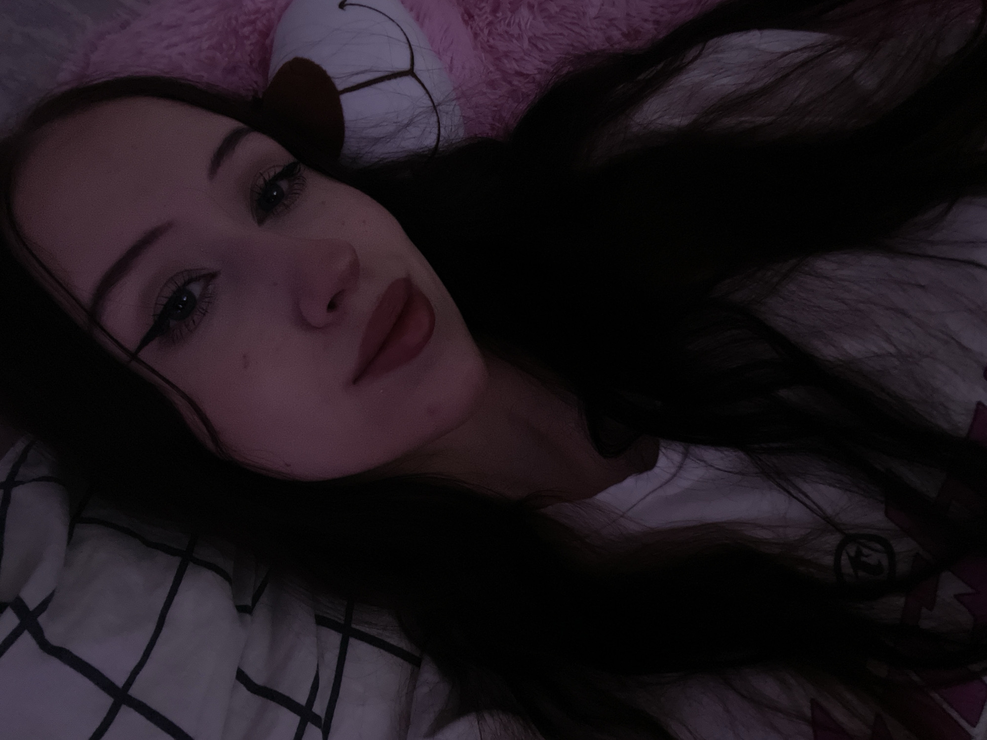 The image is a close-up, slightly angled selfie of a young woman with long, dark hair. She is lying down, with her head resting on a pillow that has a black and white checkered pattern. To her right, a large, pink, fluffy object, possibly a stuffed animal, is visible. The woman is wearing a white t-shirt with a graphic design. She has winged eyeliner and dark lipstick. The lighting is dim, casting a purple hue over the scene. There is no indication of the city of Votkinsk, Russia, or any elements that would suggest this location.