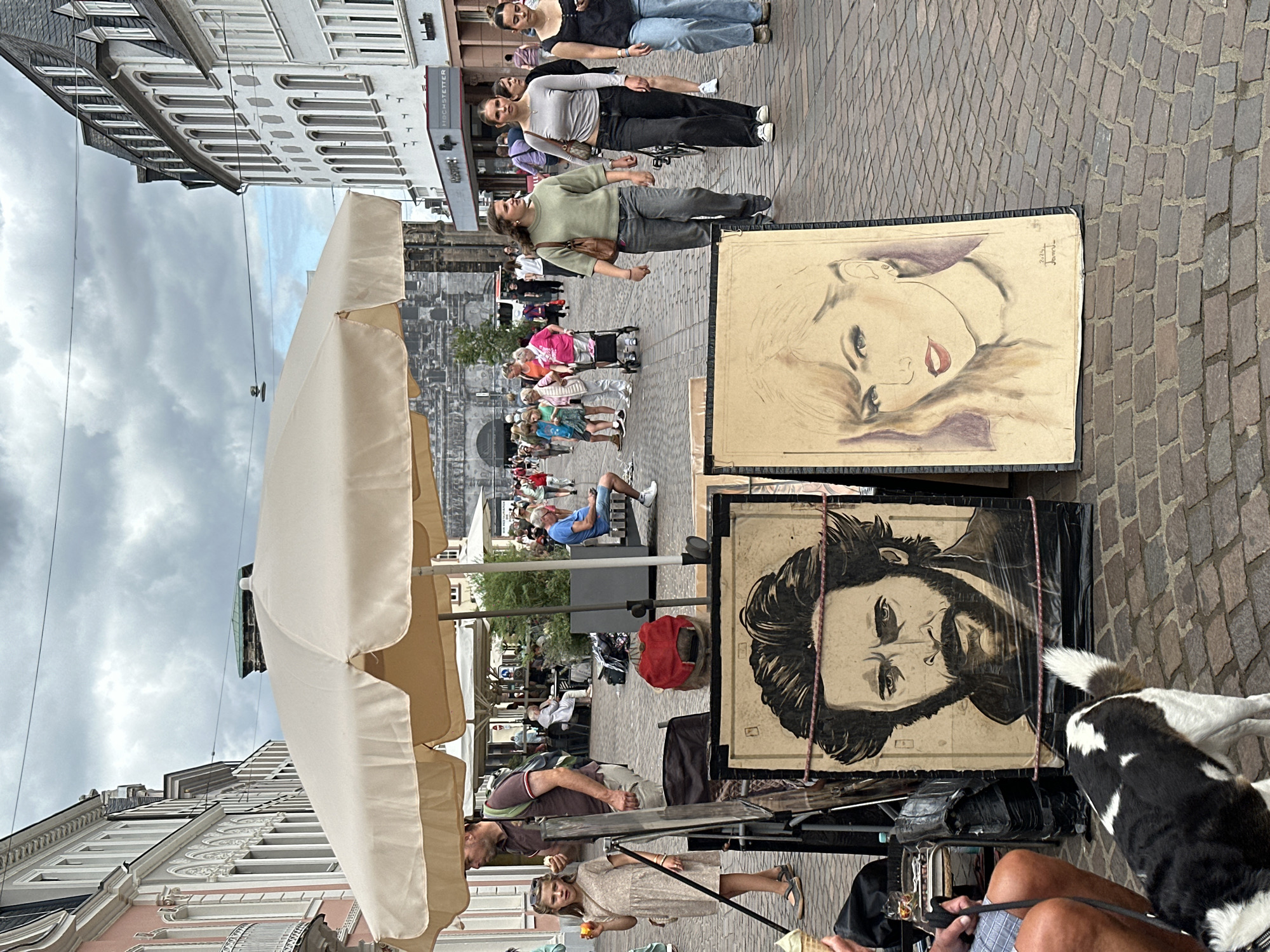 This image depicts a bustling street scene in Trier, Germany, likely a pedestrian shopping area given the architecture and presence of shops. In the foreground, two framed portraits are displayed on the cobblestone street. The portrait on the left is a detailed illustration of a man with a beard and intense gaze, while the portrait on the right is a sketch of a woman's face, identifiable as Taylor Swift.
A black and white dog stands facing away from the camera, looking towards the portraits. To the left of the dog, a person's legs are visible, wearing checkered shorts, and a leash extends from their hand towards the dog. A person in a tan dress and a man in a maroon polo shirt are standing near the display, with the man holding what appears to be a piece of fruit. A large, cream-colored umbrella is set up, providing shade.
In the background, a crowd of people strolls along the street. Some are walking, others are pushing strollers or using mobility aids. Shops with awnings line the buildings on either side of the street, with one visible sign reading "HOCHSTETTER". The architecture suggests a European city, with ornate building facades. The sky is overcast with some patches of blue. The scene captures a lively atmosphere on a day out in the city.