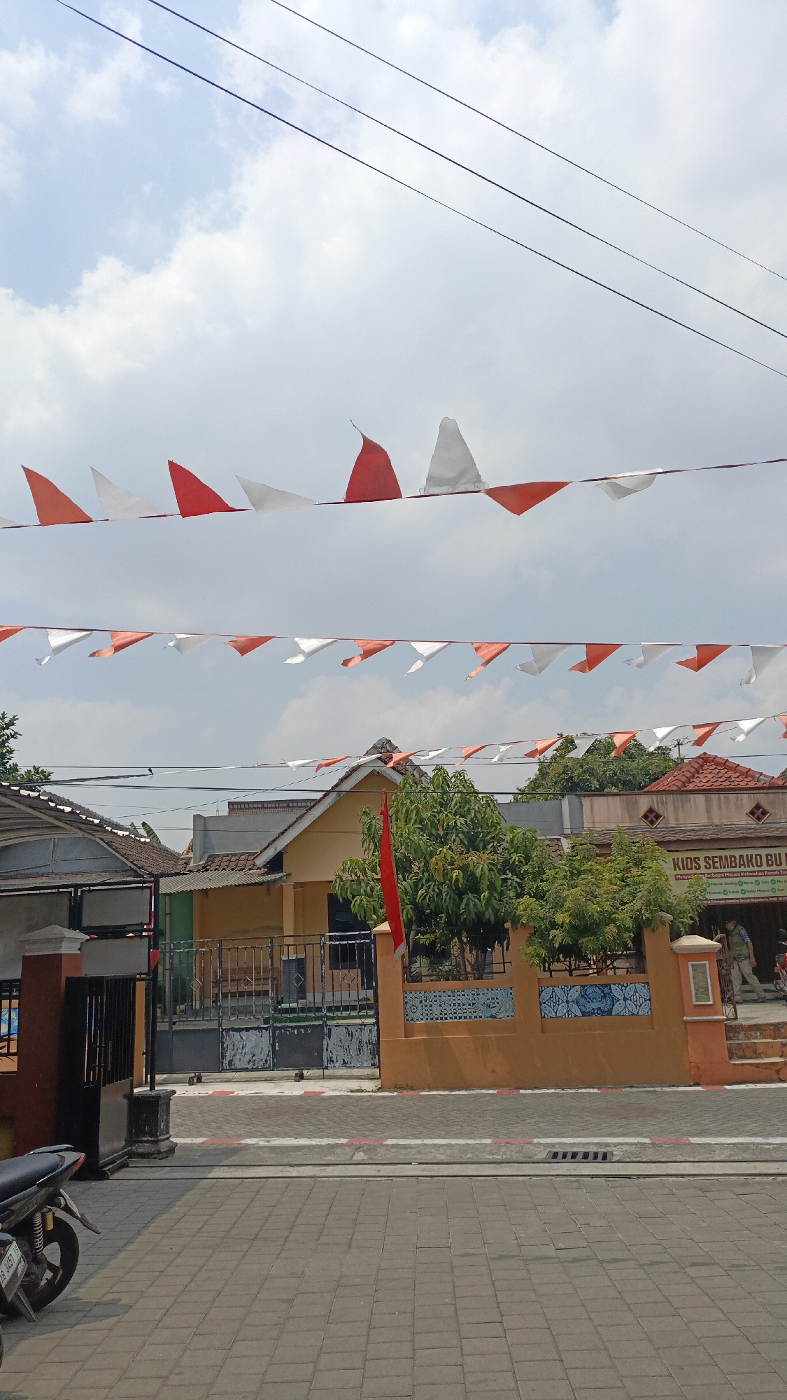 The image captures a street scene in Trihanggo, Indonesia, under a bright, partly cloudy sky. A string of red and white triangular flags is strung across the upper portion of the frame, indicative of a celebration or national holiday. Below the flags, several buildings are visible. The foreground features a paved sidewalk and street, with a motorcycle parked on the left. A fence separates the sidewalk from a building with an orange facade. The building has a sign that reads "KIDS SEMBAKO BU T..." and appears to be a small shop or business. Trees and foliage are present in front of the buildings. The overall impression is one of a typical Indonesian street with decorations for a festive occasion.