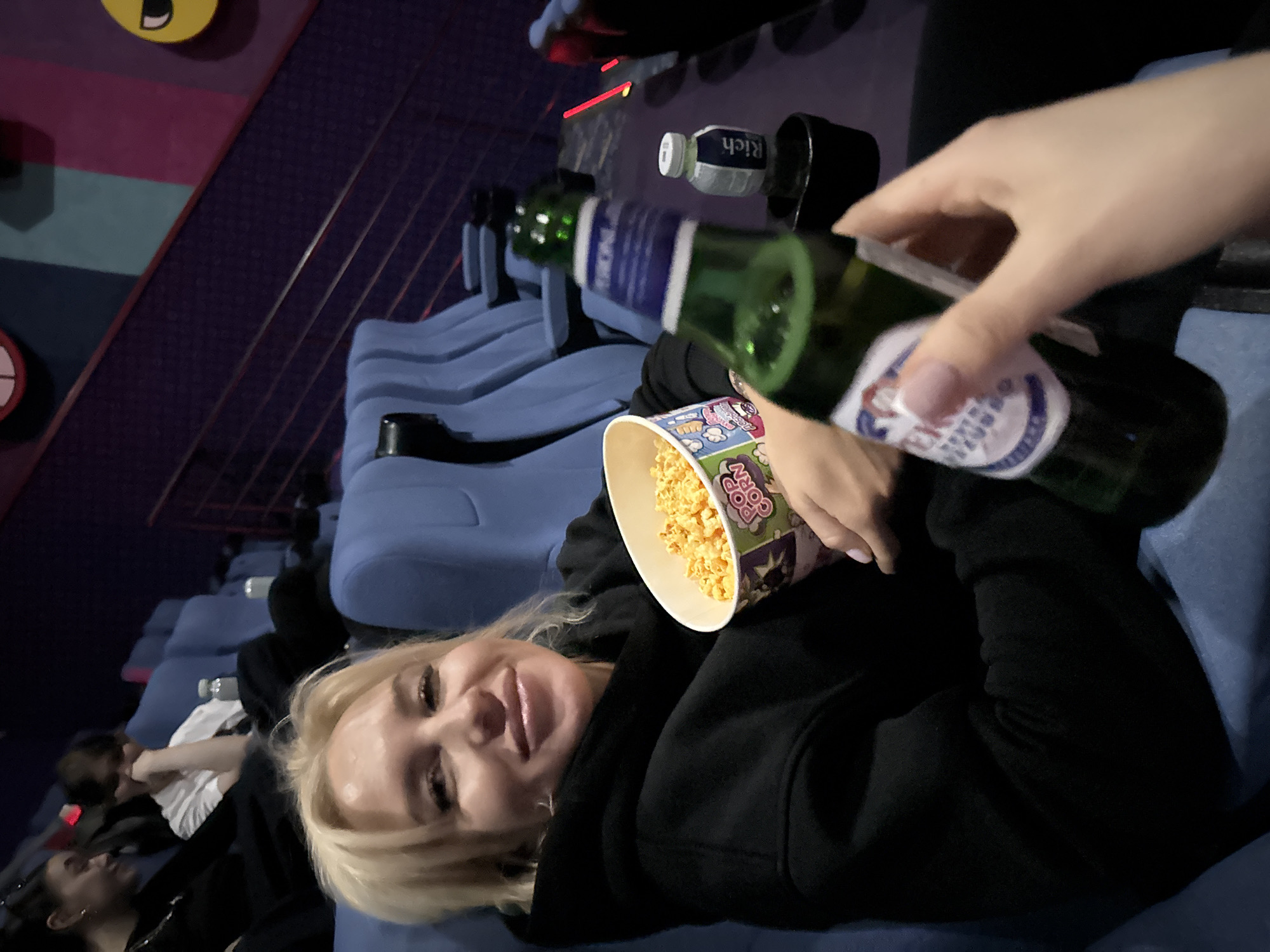 A woman with blonde hair, wearing a black hoodie, is at a movie theater. She's holding a bucket of popcorn in one hand and a bottle of beer in the other. Other moviegoers are visible in the background. The seats are blue. There are several bottles of beer lined up in the cup holders. The location is Moscow, Russia.