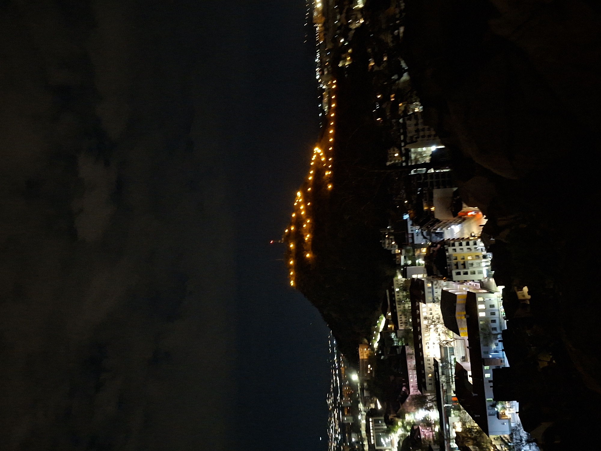 The image shows a night scene of Plovdiv, Bulgaria. The city lights twinkle below, highlighting the urban landscape. A hill, adorned with a string of lights ascending towards the top, dominates the central part of the frame. To the left, the night sky is visible, partially obscured by clouds. The right side of the image is shrouded in darkness, potentially depicting nearby foliage or structures. A cluster of buildings is evident, illuminated with varying degrees of light, possibly indicating residential or commercial areas. The lighting pattern on the hill suggests a decorative or symbolic purpose.