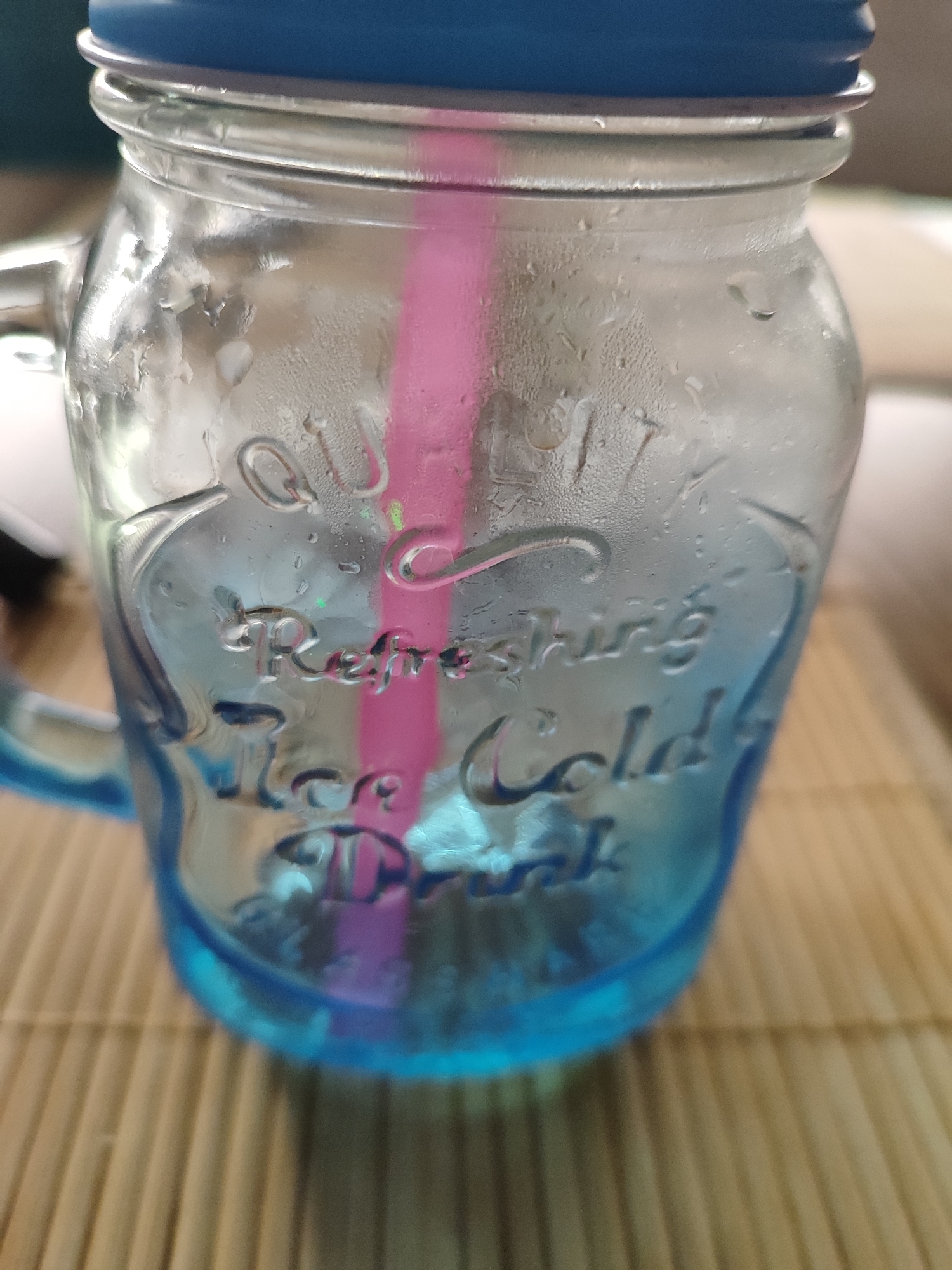 A clear glass jar with a blue lid sits on a light brown bamboo mat. The jar is filled with a light blue liquid and has condensation on the outside. A pink straw is visible inside the jar, submerged in the liquid. Embossed text on the jar reads "Refreshing Cold Drink". The background is blurred, showing a light-colored surface. The image is taken from a close-up perspective, focusing on the jar.