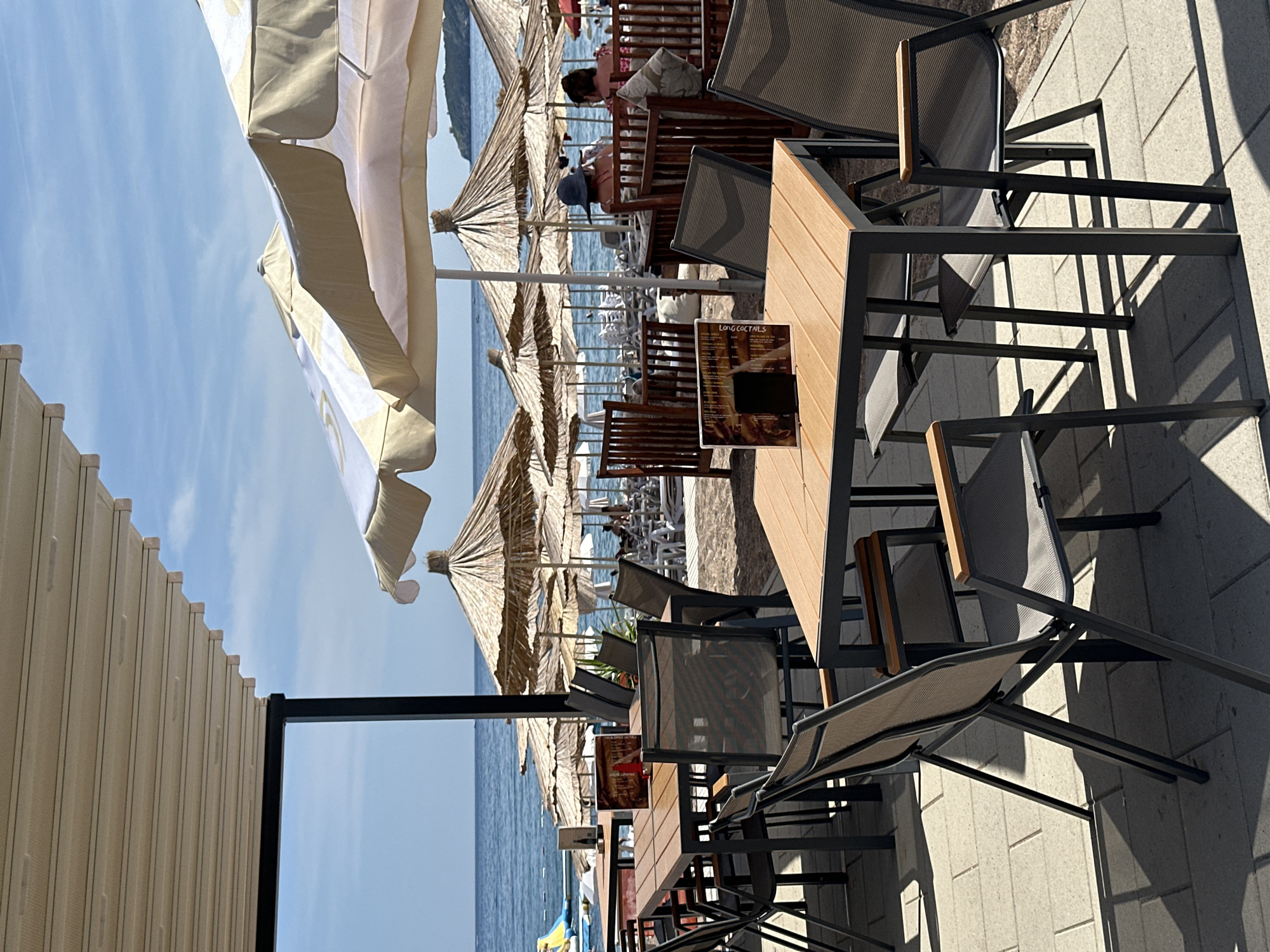 The image shows an outdoor seating area of a beachfront restaurant or cafe located in Boreti, Montenegro. In the foreground, there are several sets of tables and chairs made of dark metal frames with wooden or composite tabletops and mesh seating. A menu is visible on one of the tables.
Beyond the seating area, a sandy beach is lined with numerous straw umbrellas and lounge chairs. The Adriatic Sea is visible in the background, with calm, blue water and a clear sky. A few distant boats can be seen on the water. Some people are present on the beach, relaxing on the chairs or in the water. A water slide structure is partially visible on the left side of the frame, near the water's edge. The overall scene depicts a typical summer day at a coastal resort.