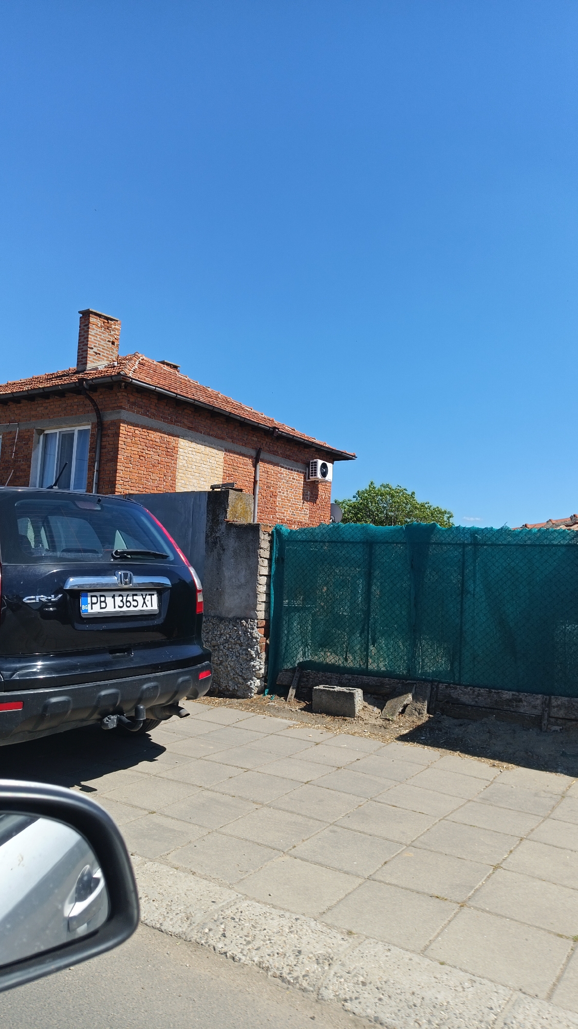 A black Honda CR-V with Bulgarian license plate PB 1365 XT is parked next to a sidewalk in Milevo, Bulgaria. A brick house is visible in the background with an air conditioning unit on the side. A green mesh fence is next to the house. The sky is clear and blue. The side mirror of another vehicle is visible in the foreground. The sidewalk is made of grey concrete slabs.