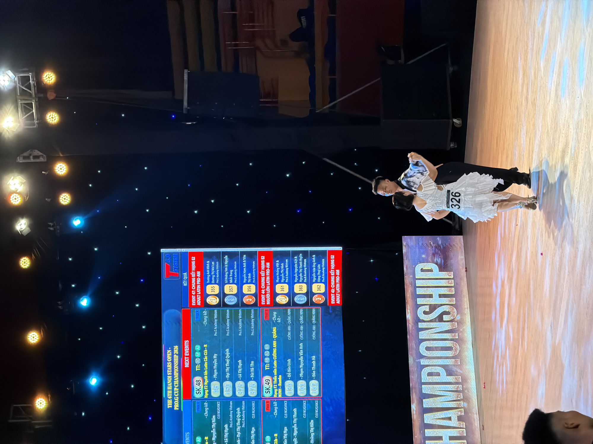 On a brightly lit stage, a couple performs ballroom dancing. The male dancer is dressed in a black shirt with a white abstract pattern and black pants. The female dancer wears a white, ruffled, short-sleeved dress. A large screen behind them displays event information and results for "THE 6TH HANOI STARS OPEN - PRO.G CUP CHAMPIONSHIP 2026". The screen shows event details including "NEXT EVENTS" and "KẾT QUẢ" (Results). The floor is illuminated with warm, orange light, and a large banner with the word "CHAMPIONSHIP" is visible at the bottom of the image. The background features a dark ceiling with numerous stage lights, some yellow and some blue, and a starry pattern. The scene appears to be indoors, likely at a dance competition venue in Hanoi, Vietnam.
