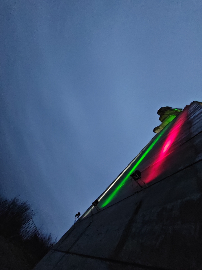 The image depicts a section of the Monument to 1300 Years of Bulgaria in Shumen, Bulgaria, observed at dusk or night. The sky occupies the upper two-thirds of the frame, exhibiting a dark, uniform blue-grey hue indicative of low light conditions. The lower right portion of the image features a large, angular concrete wall, part of the monument's brutalist architecture. This wall is illuminated by multiple artificial lights mounted directly onto its surface. A prominent green light beam extends diagonally upwards and to the right along the wall, originating from a visible light fixture. Parallel to this, a distinct red light beam also travels diagonally upwards, emanating from another visible fixture. Further to the lower left on the same wall, another green light beam and its source fixture are discernible. In the lower left corner of the frame, a dense cluster of dark, silhouetted vegetation, possibly trees or bushes, is visible against the sky, with faint outlines of a fence-like structure behind them. No individuals are present in the scene.