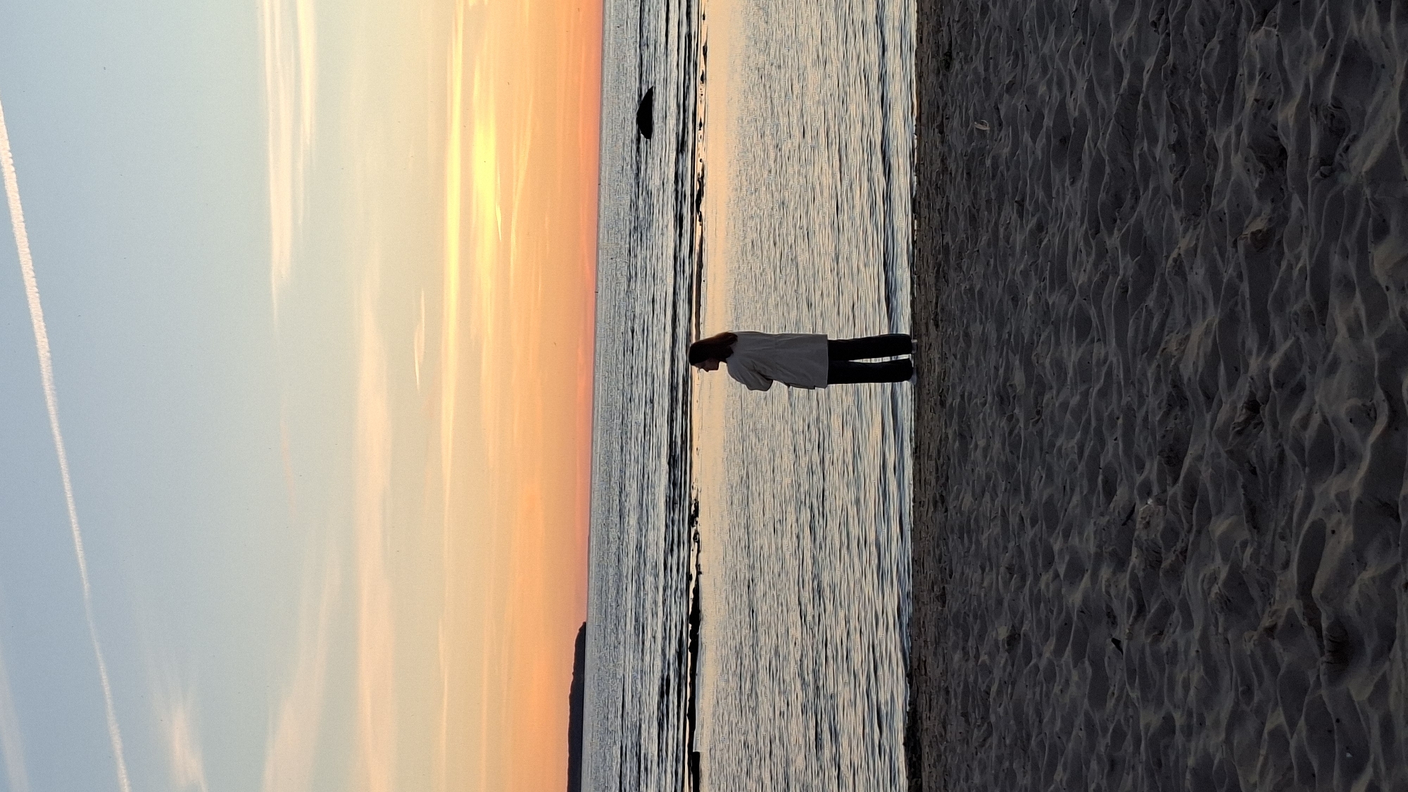 A lone figure stands on a sandy beach, facing a body of water. The sky above is a gradient of pale blue and soft orange, transitioning into a darker hue as it meets the horizon. Two white contrails are visible in the upper portion of the sky. The water's surface is calm, with gentle ripples reflecting the colors of the sunset. A faint, dark landmass can be seen in the distance on the left side of the frame, and a dark, indistinct rock juts out of the water on the right. The beach in the foreground is textured with sand dunes, casting shadows in the low light. The person is dressed in dark trousers and a light-colored jacket or shirt, with their back to the camera. Their posture suggests contemplation or observation of the seascape. The scene evokes a sense of tranquility and solitude at dusk. This is likely Kadriorg Beach in Tallinn, Estonia, known for its sunset views.