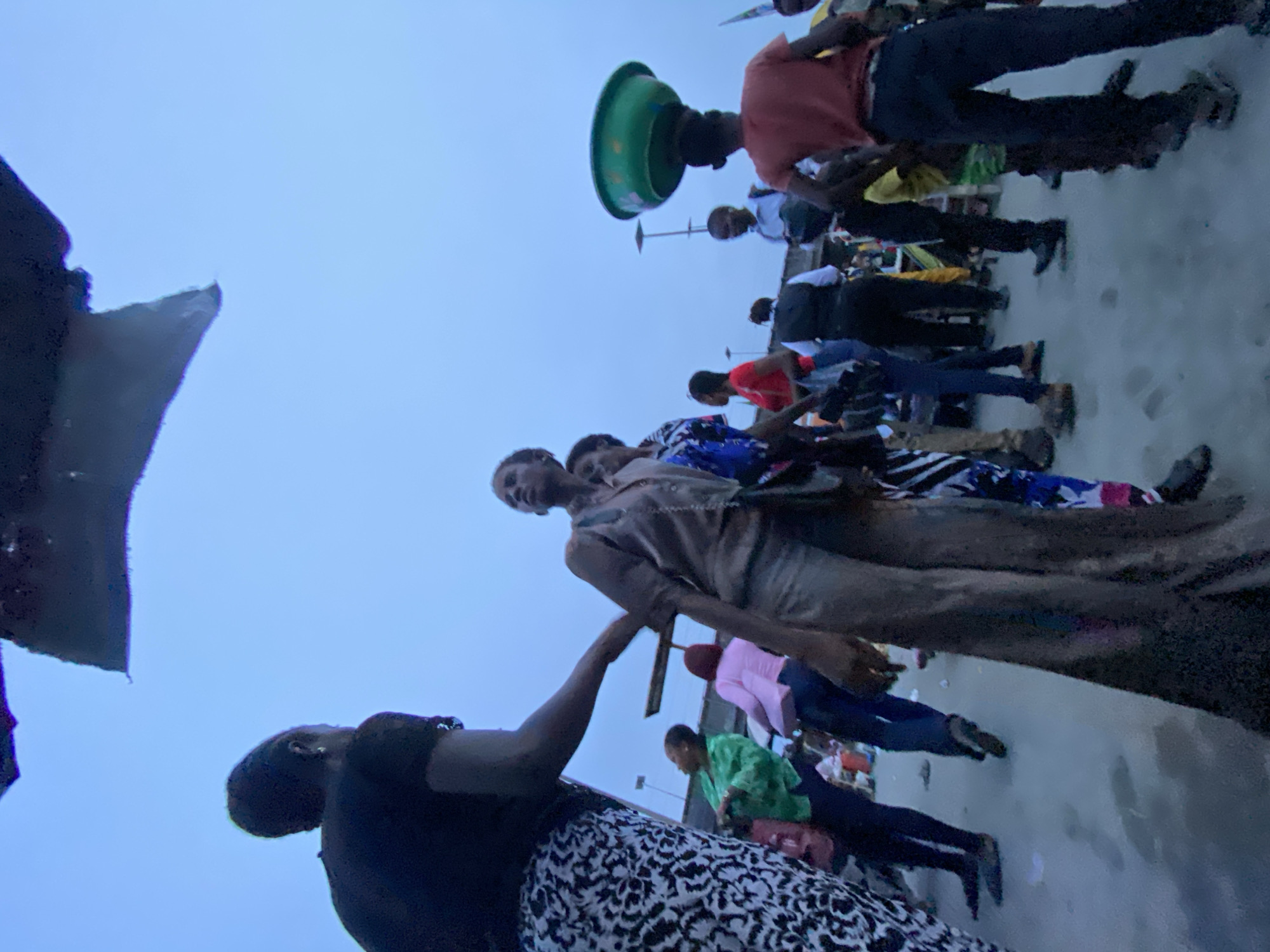A crowd of people is gathered on a concrete surface outdoors under a dim sky. A person at the forefront is wearing a black top and a black-and-white patterned skirt, reaching their hand out towards another person wearing a beige outfit. Others are standing in a line, dressed in various colors and patterns. One person further back is balancing a green bowl on their head. A black umbrella is partially visible on the left. This scene is captured in Lekki, Nigeria.