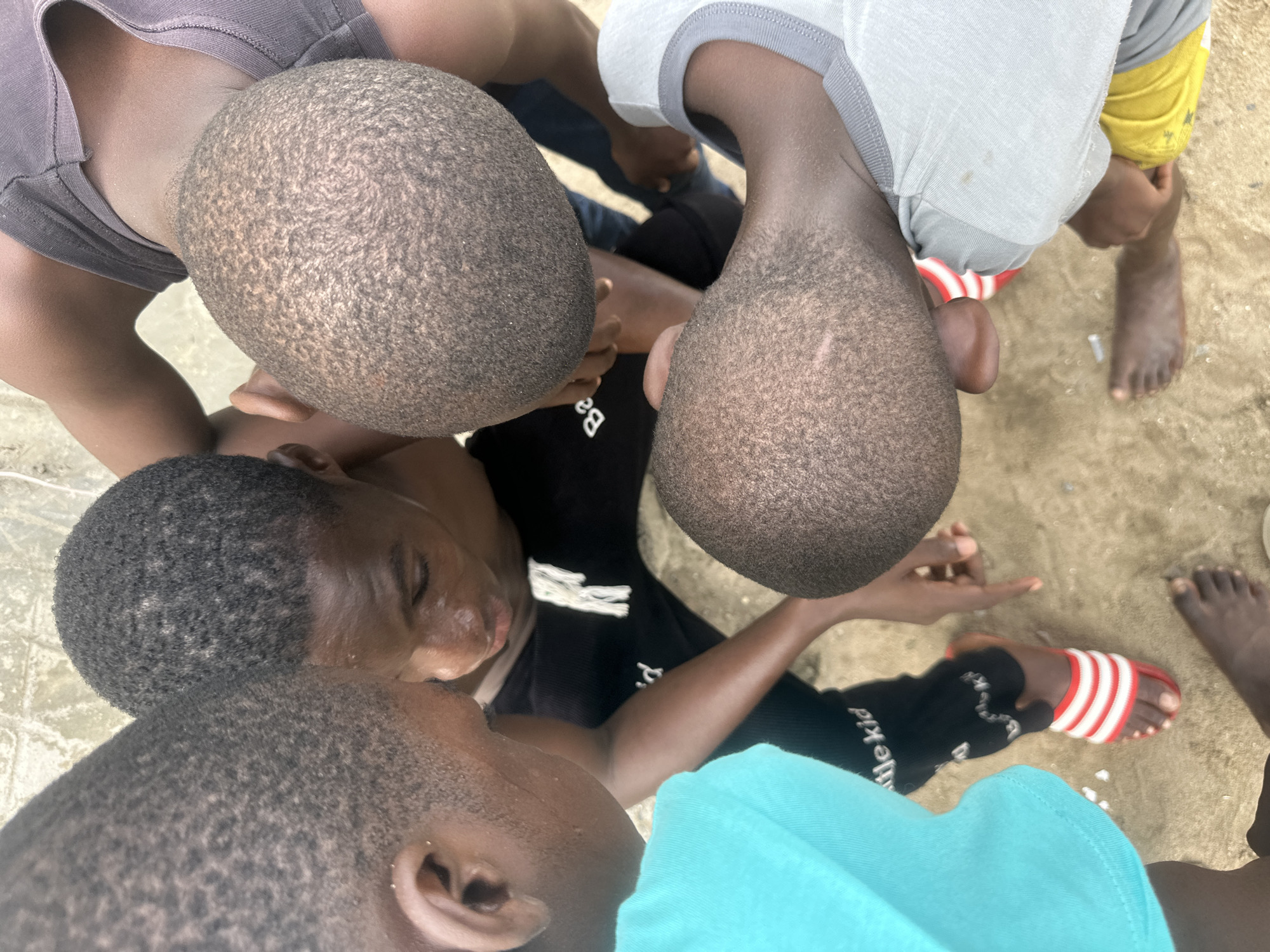 The image presents a high-angle, overhead view of a group of young individuals, likely boys or young men, gathered closely together. Their heads, covered with short, dark, textured hair, are prominent in the frame, indicating dark skin tones.
At least four individuals are discernible:
* One in the top left wears a grey t-shirt.
* Another in the top right wears a light grey t-shirt with a visible yellow sleeve. This person wears a red and white striped slide sandal on one foot, while another foot is bare on the sandy ground.
* A central figure wears a black t-shirt or long-sleeved shirt. White text is visible on the shirt, including "Ba" and "the kid". This person appears to be looking downwards into the center of the huddle, with hands seemingly engaged near the ground.
* Another individual in the bottom left wears a light blue t-shirt and is also looking towards the center of the group.
The ground beneath them appears to be sand or dirt, suggesting an outdoor setting. The individuals are positioned in a close huddle, implying a shared focus or activity, possibly looking at something on the ground or interacting closely. Their expressions are not clearly visible from this high vantage point, so specific emotions cannot be determined. The scene takes place in Badagry, Nigeria.