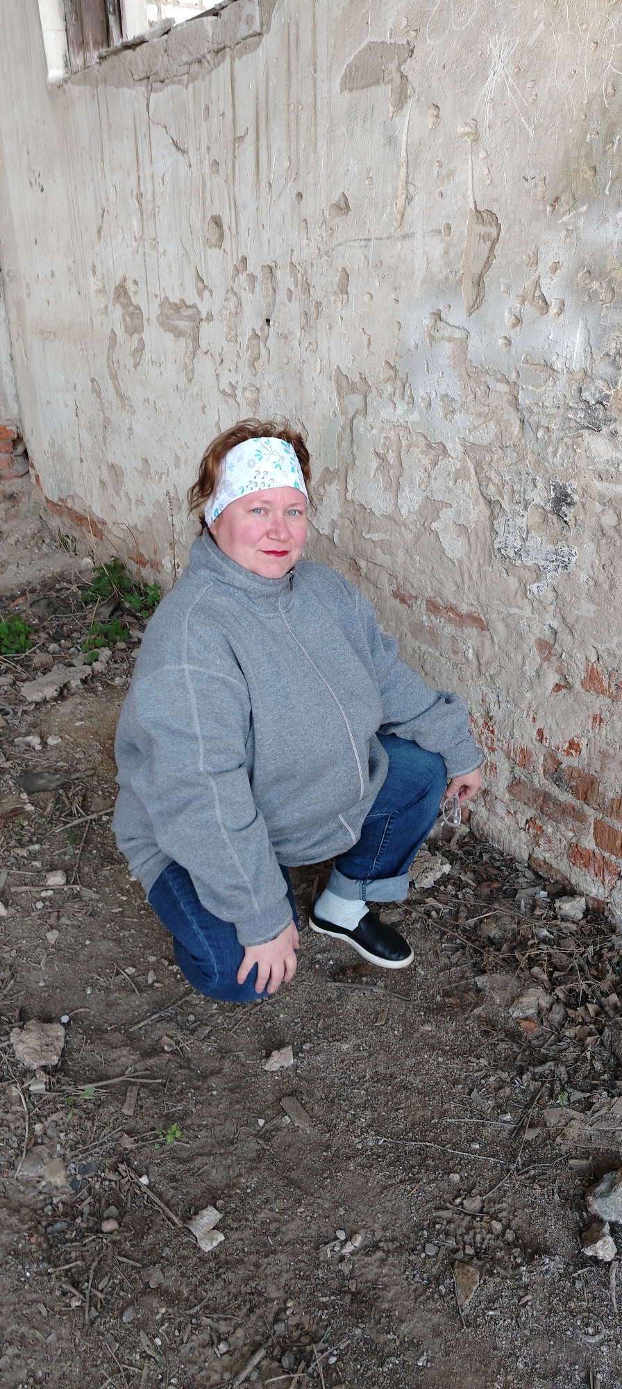 A woman is crouching near a dilapidated wall in what appears to be an outdoor setting. The woman is wearing a grey zip-up jacket, blue jeans, a white patterned headband, and black and white shoes with white socks. She has red lipstick and fair skin. The wall behind her is crumbling, with peeling plaster revealing brick underneath. The ground is covered in dirt, small rocks, and some sparse vegetation. The location is Зипуново, Russia.