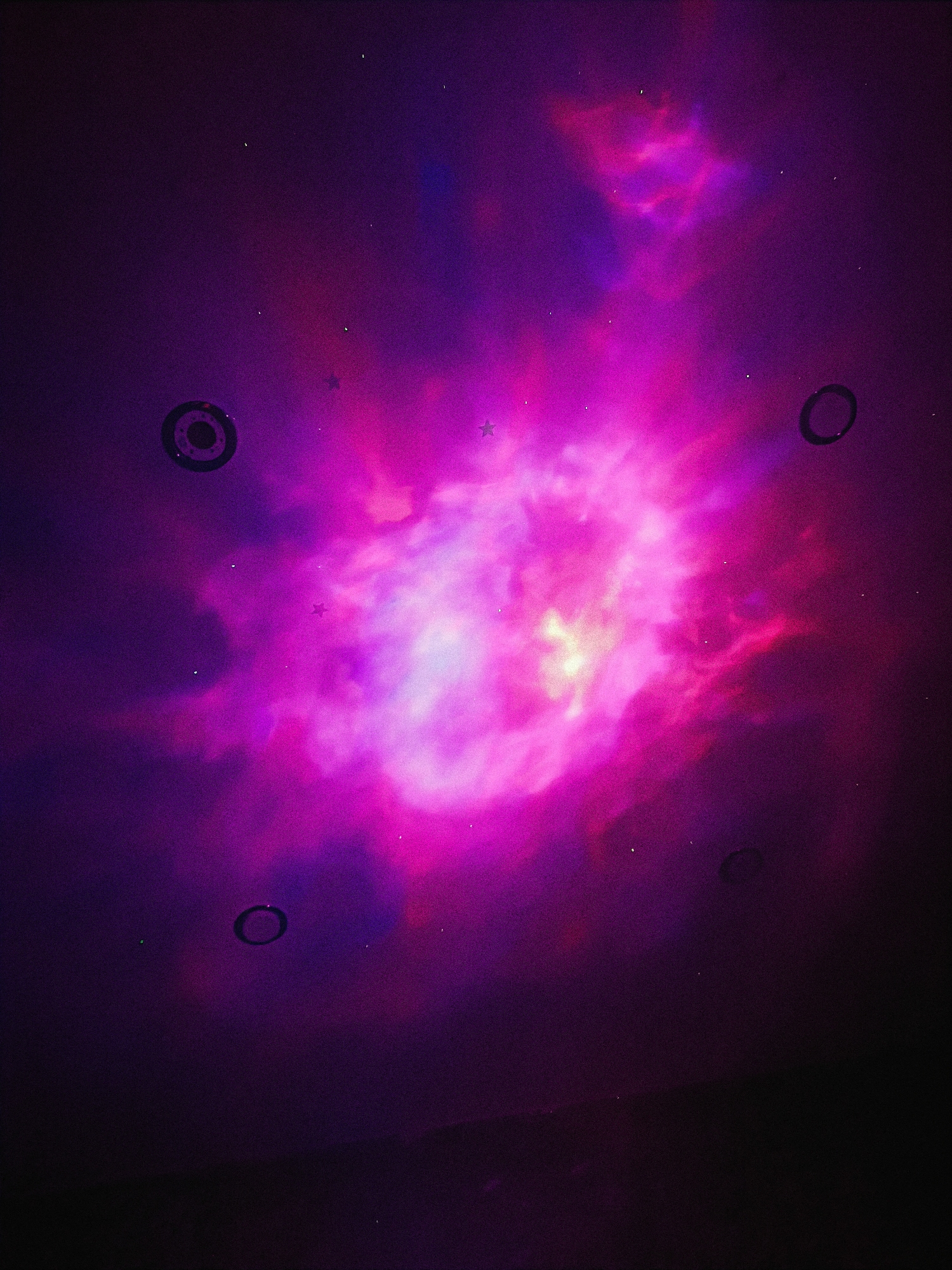 A night scene of a dark purple space with a bright, glowing nebula in the center. There are several small, black, circular objects arranged around the perimeter of the nebula. These objects appear to be physical elements in the same space. There are also a few sparse, small, star-shaped lights scattered across the nebula. The bottom of the image has a dark, solid surface. The specific location in Chaikovsky, Russia is not discernible from the image.