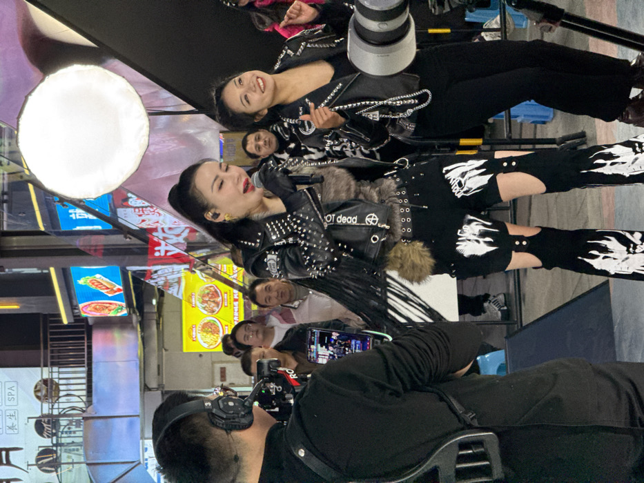 The image depicts an outdoor night scene, likely a live performance or broadcast event, in the Yuzhong District of China.
In the foreground, a female performer with dark hair pulled into a high ponytail is centrally positioned, holding a microphone to her mouth, suggesting she is singing or speaking. She wears a black studded leather jacket with long fringes on the sleeves, a faux fur vest, black fingerless gloves, large gold earrings, black shorts, and black pants with thigh cut-outs and white flame-like designs on the lower legs. To her immediate right, a younger female stands smiling and looking towards the performer. This individual wears a black studded leather jacket over a black top, black pants, and has a silver-colored chain draped over her left shoulder.
To the left, a male cameraman is visible from behind, wearing a black long-sleeved shirt and black over-ear headphones with a boom microphone. He operates a professional video camera mounted on a rig, with a small monitor displaying what appears to be live footage.
In the mid-ground, several individuals, predominantly adult males, are visible as an audience, observing the event. The background features a vibrant urban environment with multiple brightly illuminated commercial signs displaying Chinese characters, food imagery (including hot dogs and noodles), and the English word "SPA." A large, circular white LED light panel is positioned to the upper right, serving as a primary light source for the scene. To the right of the smiling female, a large silver-colored cylindrical object, possibly a camera lens or lighting equipment, is visible on a tripod. A black and white geometric structure is partially visible behind and between the central figures. The scene is artificially lit, indicating an evening or nighttime setting.
