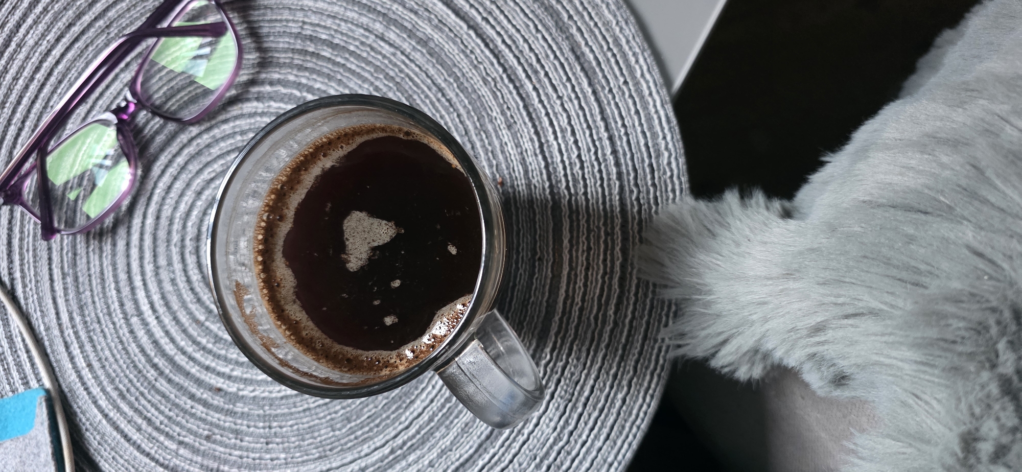 A top-down shot shows a clear glass mug of coffee with crema on a gray, circular placemat. To the upper right, a pair of purple-rimmed glasses rests on the placemat. The lower portion of the image features a soft, light gray furry object. The scene is set indoors, but no specific location in Arun, United Kingdom can be determined. There are no people or actions depicted.