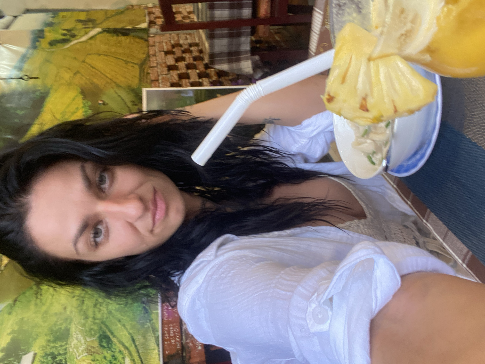 A woman with long, dark hair and fair skin, wearing a white shirt, is photographed from a slightly elevated, canted perspective. She is looking at the camera with a neutral expression. A partially consumed beverage, consisting of a pineapple in a plate with what appears to be some cream is positioned in front of her, accompanied by a white plastic straw.
The setting suggests an outdoor cafe or restaurant in Phu Thuy Ward, Vietnam. Visible in the background are portions of outdoor decorations or artwork depicting landscapes and brick patterns. A wooden chair with a plaid seat is also visible.
