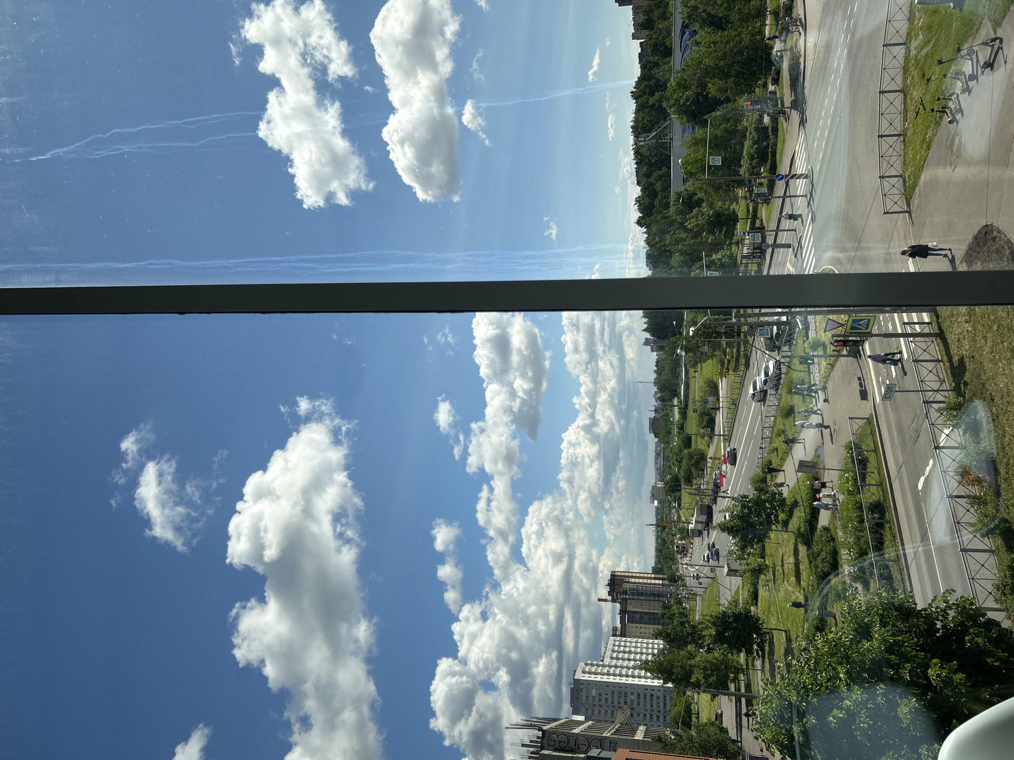 The image captures a street view from a high vantage point, likely from inside a building, looking out onto a city street in Saint Petersburg, Russia. The sky is a bright blue with scattered white cumulus clouds. A dark vertical frame, presumably a window frame, is visible on the right side of the image.
In the foreground, a wide, multi-lane road curves to the right. Pedestrians can be seen walking on the sidewalks and crossing the street at marked crosswalks. Several vehicles, including cars and a truck, are driving on the road. A row of parked electric scooters is visible on the grassy area to the right.
Beyond the road, there is a green park-like area with trees and grass. In the middle ground, a line of modern apartment buildings and other structures are visible, some with visible construction scaffolding. Further in the background, a dense line of trees forms a natural boundary, and beyond that, more buildings and structures of the city can be seen stretching towards the horizon under the bright sky. The overall scene depicts a typical urban environment on a clear day.