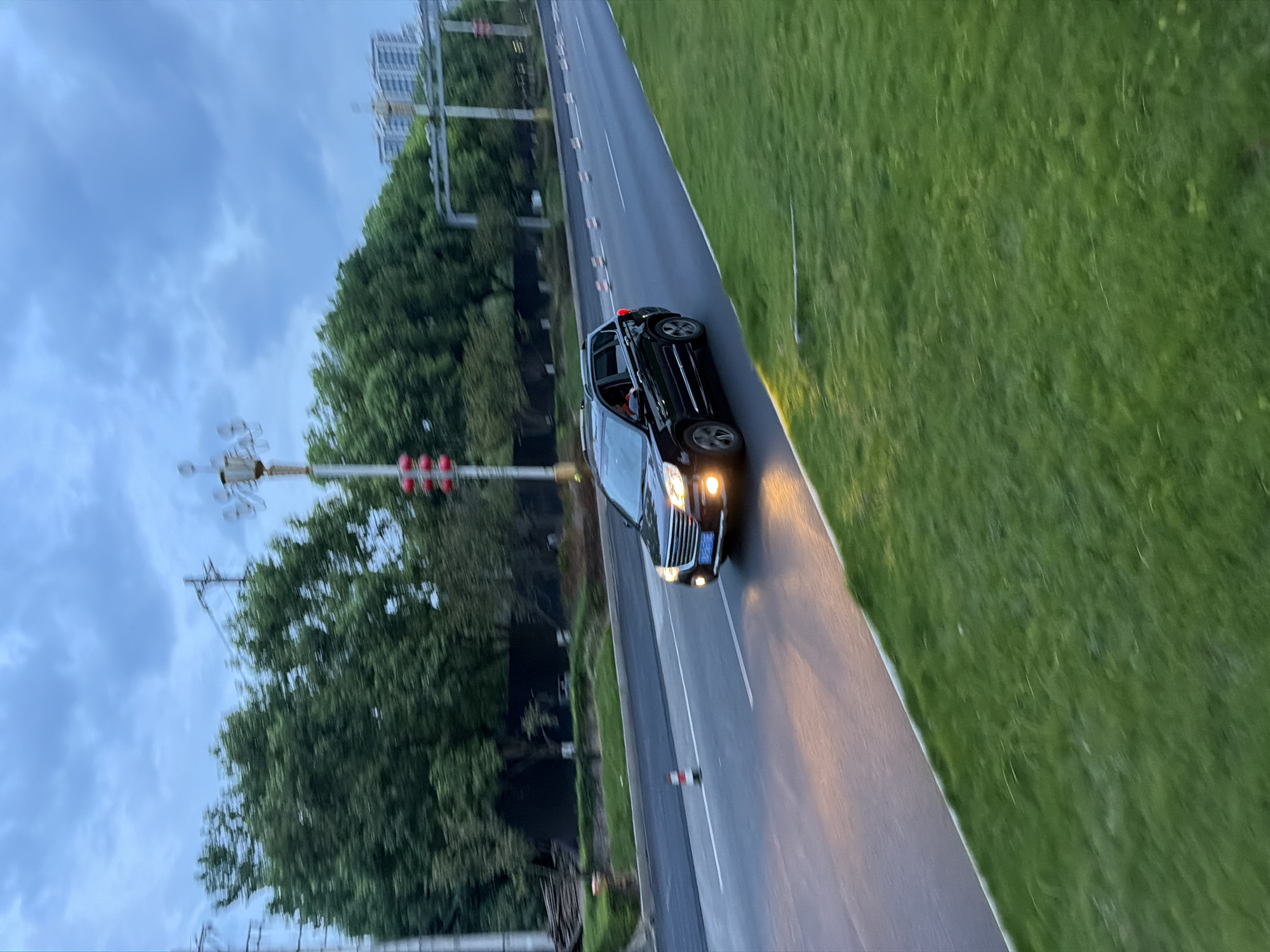 A black SUV is captured in motion on a road in Jiangjin, China. The vehicle is traveling on the right side of the road, with its headlights illuminated. The setting appears to be an urban environment with trees lining the road and buildings visible in the distance. The sky is overcast with clouds, suggesting it may be early evening or a cloudy day. The road is paved and has lane markings. There is a grassy area on the right side of the road, and traffic cones are visible on the left side of the road. The image is slightly motion-blurred, indicating movement of either the camera or the vehicle.