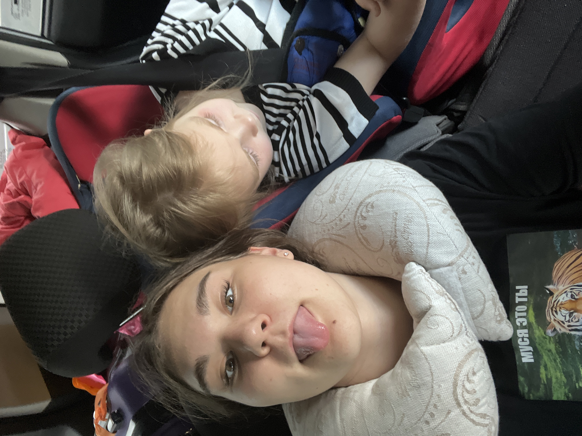 The image shows two individuals, likely related, inside what appears to be a vehicle. The foreground features a young woman with her tongue sticking out, wearing a black t-shirt with a tiger graphic and the Russian text "МУСЯ ЭТО ТЫ" which translates to "Musya, is that you?". She has a travel pillow around her neck. Behind her and to the right, a young child is visible, leaning against a car seat and wearing a black and white striped shirt. The child's head is tilted down and they appear to be looking at something out of frame. Various bags and car interior elements are visible in the background, suggesting a journey. The scene is lit by ambient light, consistent with being inside a vehicle during the day. There is no visual information within the image to suggest the location is the city of Goldyrevskii, Russia.