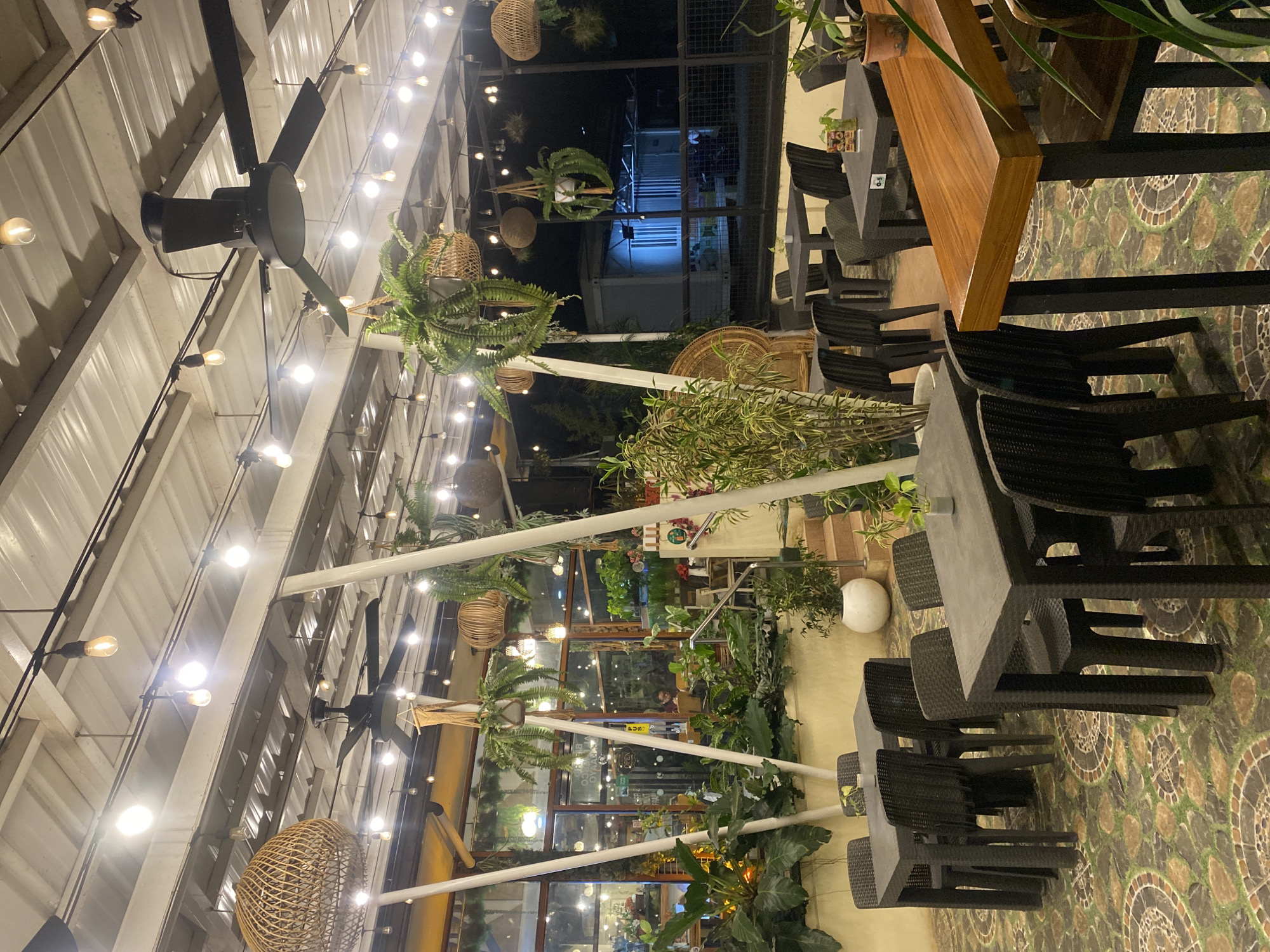 The image depicts an outdoor dining area with a corrugated metal roof supported by white posts. The space is illuminated by numerous Edison-style bulbs hanging from wires, creating a warm glow. Several black ceiling fans are visible, mounted on the roof structure. Woven wicker lampshades and hanging planters with lush green ferns are strategically placed throughout the area, adding a natural and bohemian ambiance.
The dining area features multiple tables and chairs. Some tables are square and made of dark plastic or rattan material, with matching chairs. Another table in the foreground is a rectangular wooden bar-style table with black legs, accompanied by high chairs. The floor is paved with a decorative mosaic tile pattern in earthy tones.
In the background, through large glass windows and doors, the interior of what appears to be a restaurant or cafe is partially visible. A few patrons can be seen seated inside. The exterior surroundings show glimpses of trees and a darker night sky. The overall scene suggests a relaxed and inviting outdoor seating area for a restaurant or cafe, likely in a tropical climate given the abundant plant life. While the specific location is not explicitly stated in the image, the mention of Davao City, Philippines, would imply this is an establishment within that urban area, known for its tropical environment and vibrant culinary scene.