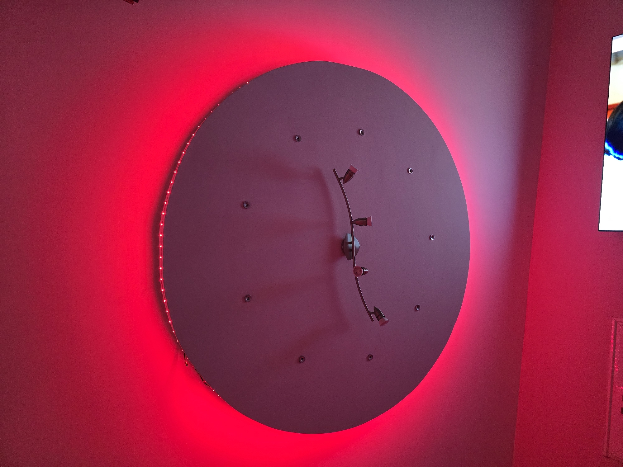 The image shows a circular object mounted on a wall, lit with red LED lights around its perimeter, casting a red glow on the wall around it. The object appears to be a light fixture, with smaller spotlights attached to a central support. There are several screws or fasteners visible on the surface of the object. Part of a television screen is visible on the right side of the image, indicating an interior setting. The lighting and color scheme dominate the scene, obscuring further details. The location, Burgas, Bulgaria is not discernible from the image.