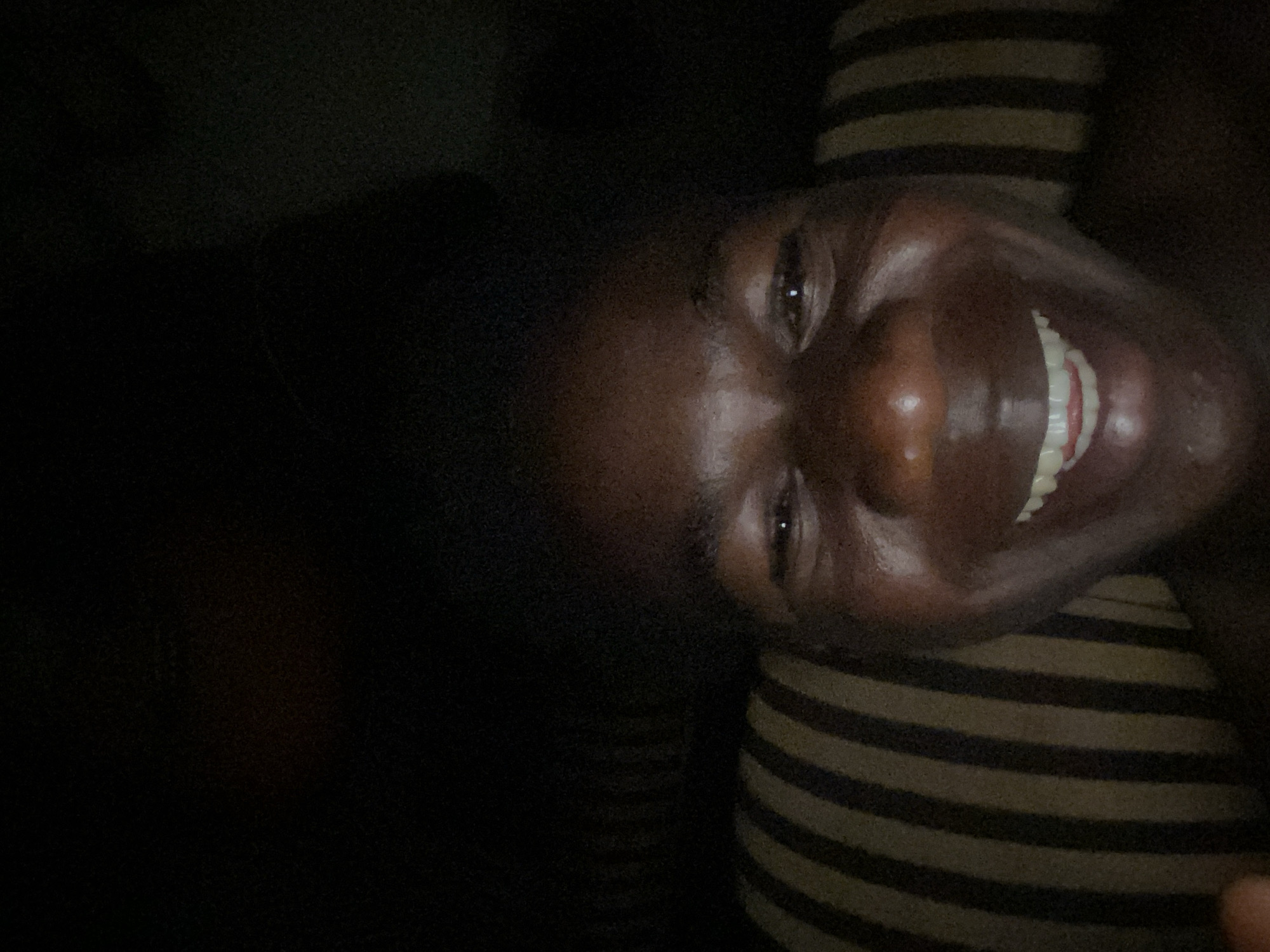 A close-up, low-light photograph depicts a Black person with a wide smile, revealing their teeth. They are positioned on a surface with horizontal, brown and black stripes. The left side of the image is largely dark and undefined. The person's face is well-lit enough to discern features like eyes, nose, and the reflection of light on their skin. The photograph appears to have been taken at a close range, filling much of the frame with the individual's face.
Location information is not discernible from the image.