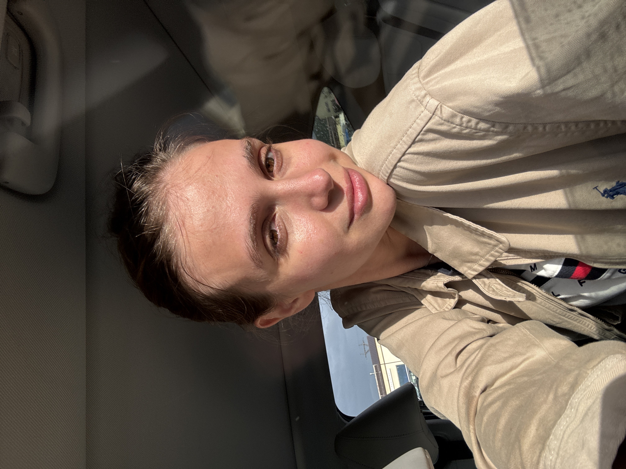 A woman is taking a selfie inside a vehicle. She is wearing a tan jacket, possibly from Ralph Lauren, over a Tommy Hilfiger shirt. The surrounding environment includes a car interior, a window showing a building and sky, and based on other visual data, the probable location is Bagratashen, Armenia.