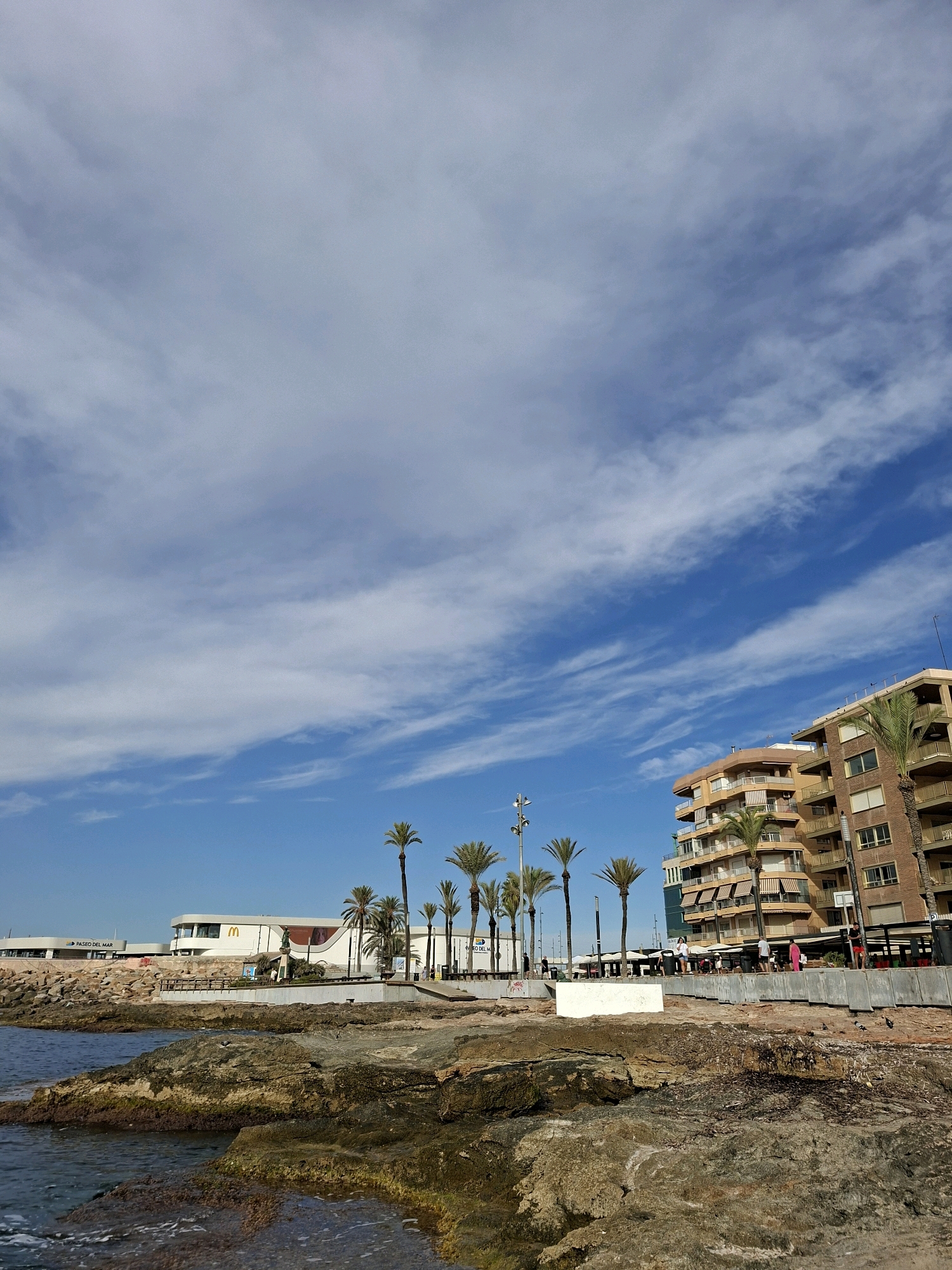 The image depicts a coastal scene in Torrevieja, Spain, with a view of the Mediterranean Sea on the left and a promenade with buildings and palm trees on the right. The foreground features a rocky shoreline with some vegetation and the sea. In the mid-ground, there is a paved promenade with several people walking and standing. Along the promenade, there are various structures, including a McDonald's restaurant with its iconic golden arches logo, a modern white building with large windows, and a multi-story residential building with balconies. Several tall palm trees are interspersed along the promenade, adding to the tropical ambiance. The background is dominated by a bright blue sky with scattered white clouds. The lighting suggests it is daytime, likely mid-morning or afternoon, with sunlight illuminating the scene.