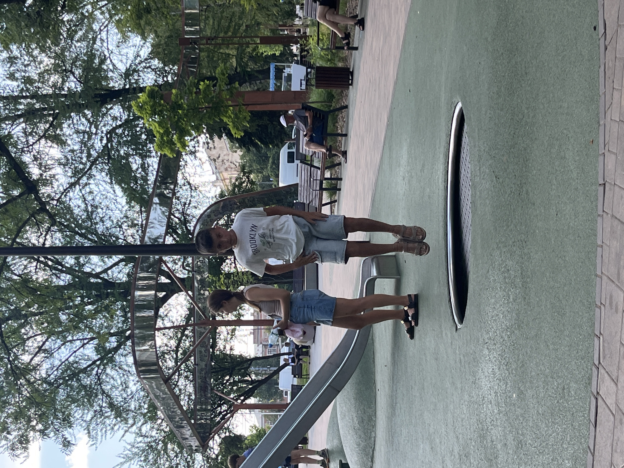 A boy and a girl stand on a green play area. The boy is on the left, wearing a white t-shirt with "BROOKLYN" printed on it and blue shorts. The girl is to his right, wearing a light-colored tank top and a blue denim skirt. Both are wearing sandals. They appear to be standing on a slide structure. In the background, there are trees with lush green foliage and a cloudy sky, suggesting it's daytime with possibly overcast weather. There are also park benches and a trash can visible, hinting at a public park setting. A large, circular, reflective structure, possibly an art installation or part of the playground, is partially visible behind them. The overall scene suggests a casual outdoor activity, likely in a city park. The presence of people and playground equipment indicates a recreational area. The orientation of the image is sideways.