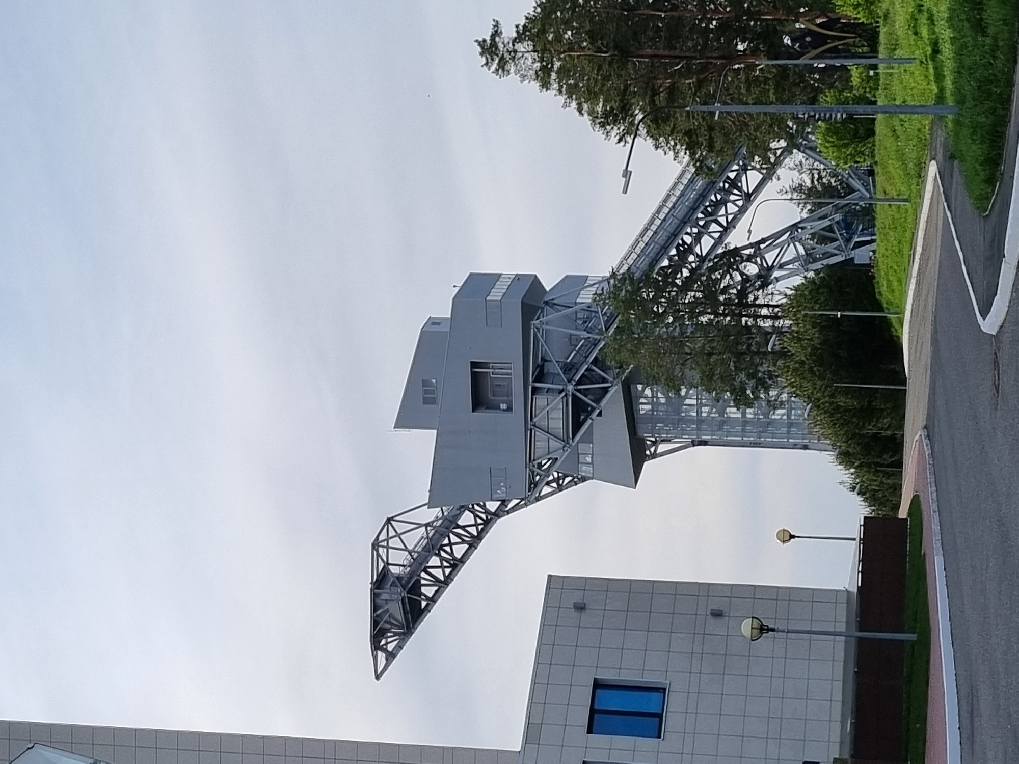 The image shows the observation tower in Прикамский, Russia. The tower is a steel structure with a distinctive angular design. It has a viewing platform at the top, accessible by a series of stairs and ramps. In the background, there are trees and a building with a blue window. The sky is overcast. A street and sidewalks are visible.