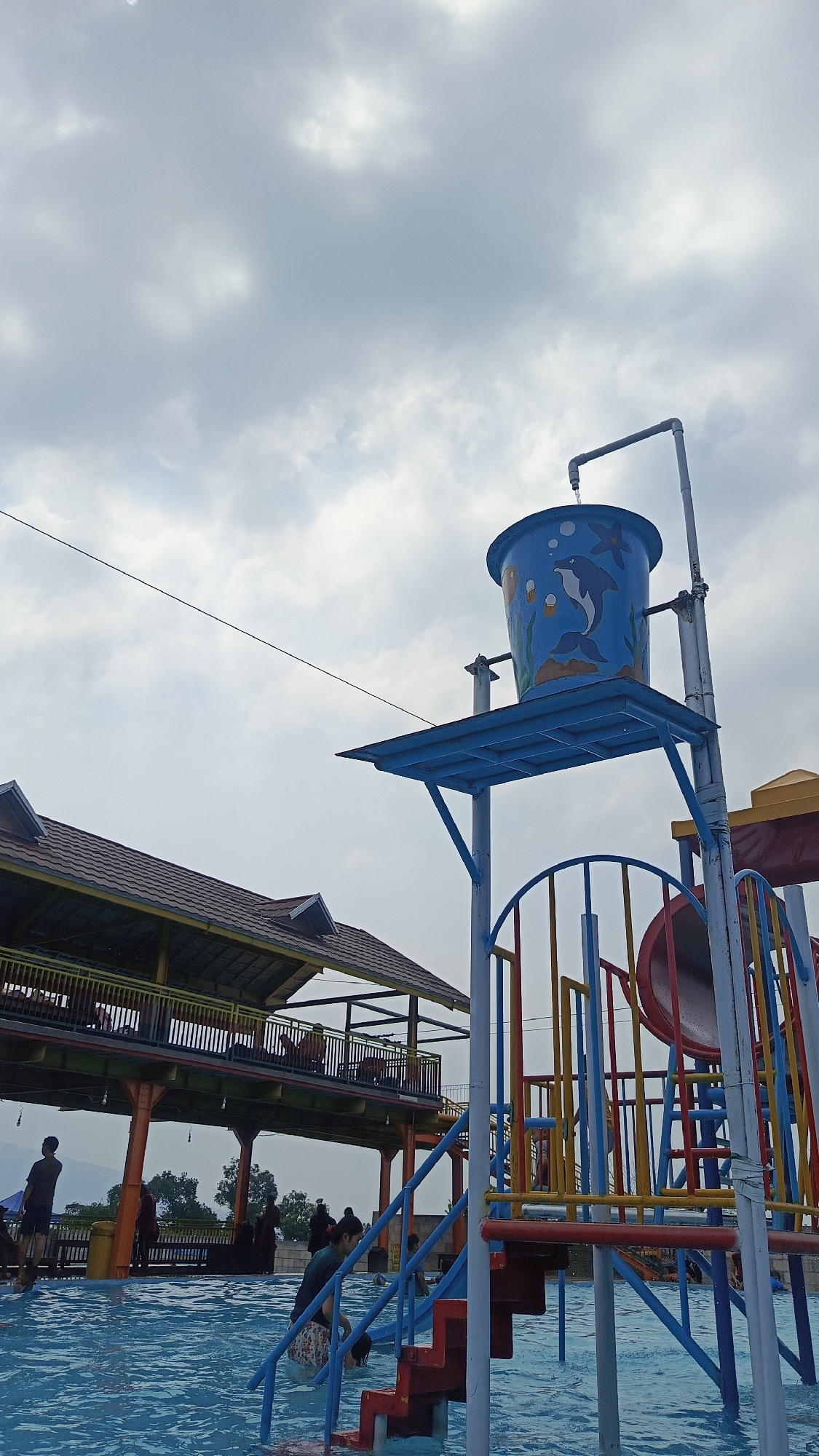 The scene is a water park in Prigen, Indonesia. A large, blue bucket decorated with marine life, including dolphins and starfish, sits atop a blue metal structure with multiple slides and ladders in primary colors. Water drips from a pipe into the bucket. The structure is partially submerged in a pool of water.
In the background is a two-story structure with a brown roof, yellow trim, and orange support columns. People are visible on both levels. In the pool, several people are visible engaging in recreational activities. The sky is overcast with light gray clouds. A thin cable stretches across the upper part of the image.