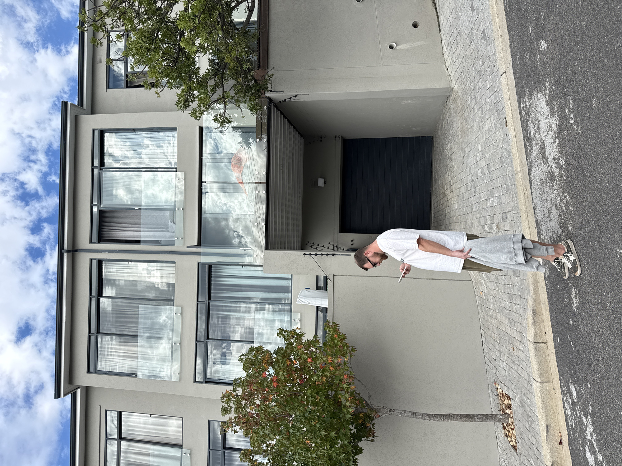 A man wearing a white t-shirt, gray shorts, and sneakers stands on a paved sidewalk next to a gray building. He holds a smartphone in his hand, looking down at it. The building has multiple stories, with large windows and glass balconies. A tree with green and reddish leaves is in front of the building. Above the man and the sidewalk is an entrance to what appears to be a garage with a closed black door. The sky is visible in the upper left corner, showing blue sky and some white clouds. The location appears to be an urban setting in Cape Town, South Africa.