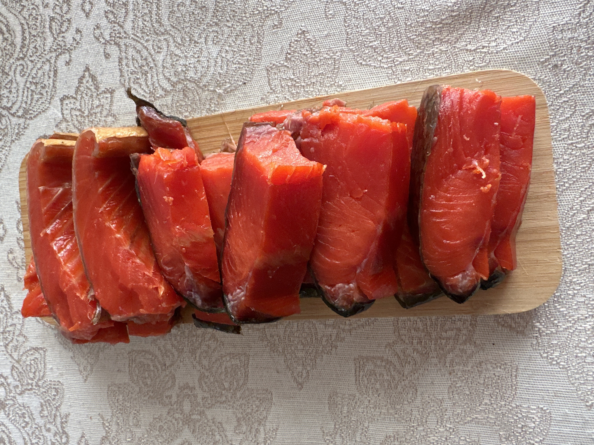 A top-down, eye-level shot shows a wooden cutting board on a textured tablecloth. The cutting board, positioned diagonally from top-left to bottom-right of the frame, is laden with approximately ten bright red, smoked salmon fillets. The fillets are arranged in a slightly overlapping, stacked manner, filling the majority of the board. The tablecloth has a repeating floral pattern in a light beige color. The scene is lit with natural light, casting subtle shadows and highlighting the glistening texture of the fish. The image captures a close-up view of the food, with no people or other objects visible. The location is not specified in the image itself, but assuming it is in Khabarovsk, Russia, it would be a domestic setting, possibly a kitchen or dining room.