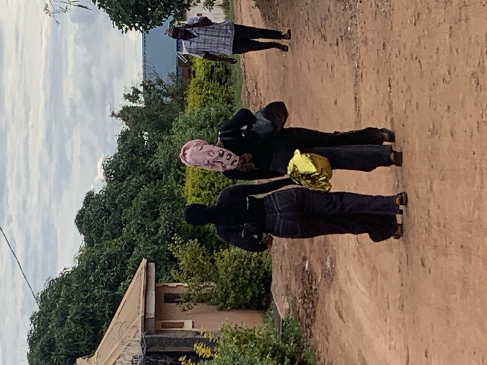 This image captures a daytime scene on an unpaved, reddish-brown dirt road in Jos, Nigeria, under an overcast sky with white and grey clouds.
Three individuals are visible on the road. In the midground, two females are walking away from the camera. The leftmost female wears a black long-sleeved top, dark denim jeans, and dark sandals, carrying a small black bag over her left shoulder. Her head is covered by a dark fabric. The female to her right wears a black long-sleeved top, dark trousers, and dark sandals. Her head is covered by a pink patterned fabric. She carries a dark shoulder bag on her left side and a crumpled gold-colored metallic object in her left hand. Further down the road on the right, a male figure is walking towards the right side of the frame. He wears a light-colored plaid shirt, dark trousers, and a patterned cap, holding a dark object, consistent with a mobile phone, to his right ear. He also wears dark sandals.
To the left, a portion of a single-story building with a light-colored wall and a brown corrugated roof is visible, partially obscured by dense green foliage. More dense green vegetation, consisting of trees and bushes, dominates the background on both sides, with a distant blue-roofed building partially visible through the foliage on the right side. A thin dark line, possibly a utility cable, crosses the upper left portion of the sky.