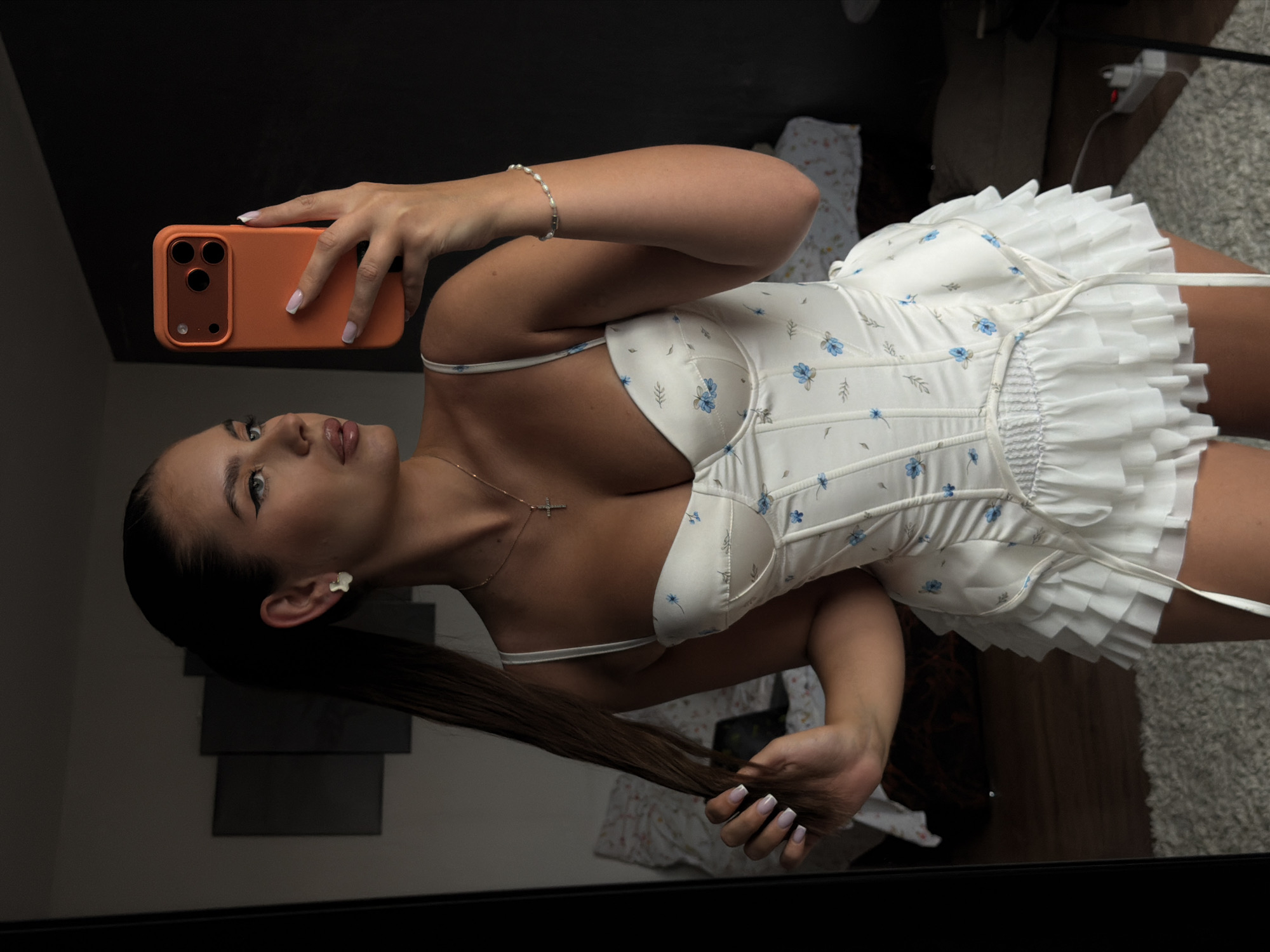 A young woman is taking a selfie in front of a mirror. She is holding an orange iPhone in her left hand, with her right hand grasping the end of her long brown hair. She is wearing a white floral dress with a corset top and a layered ruffle skirt. She also has a bracelet on her left wrist and a necklace with a cross pendant. The background appears to be a bedroom with a bed and other furniture. It is unlikely this image was taken in Krasnoyarsk, Russia.