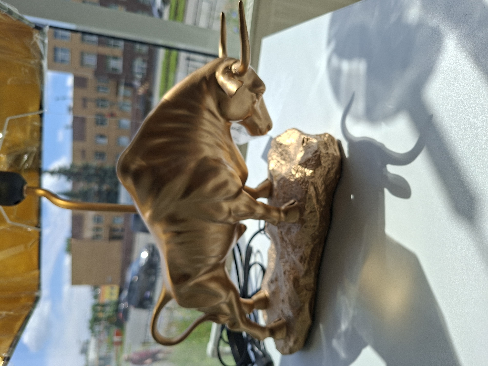 An indoor scene shows a golden bull figurine on a white surface, casting a shadow. The figurine is made of metal and depicts a bull with its head lowered and horns extended. The background is blurred, showing buildings, trees, and a cloudy sky, indicative of an urban environment, possibly Izhevsk, Russia, given the context. A yellow lampshade is visible at the top of the frame, suggesting the figurine might be part of a lamp. The lighting suggests daytime with sunlight streaming in, creating distinct shadows. No individuals are clearly discernible or interacting with the figurine.