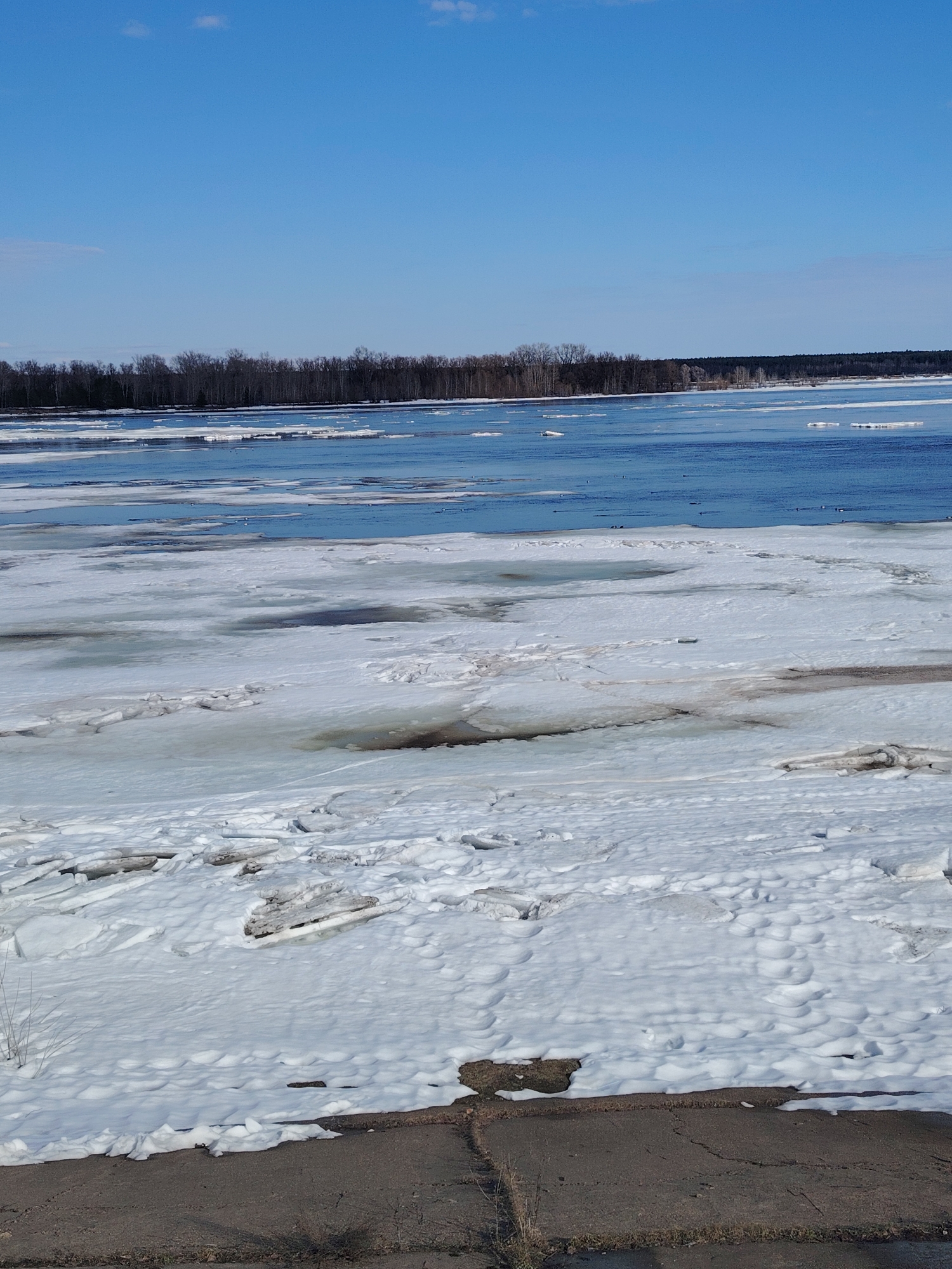 The image depicts a partially frozen river landscape. In the foreground, there is a layer of ice and snow covering the ground and extending onto the water. Patches of open water are visible among the ice floes. In the distance, a line of trees marks the opposite bank of the river. The sky is blue with some scattered clouds. The location is near the city of Sarapul, Russia.