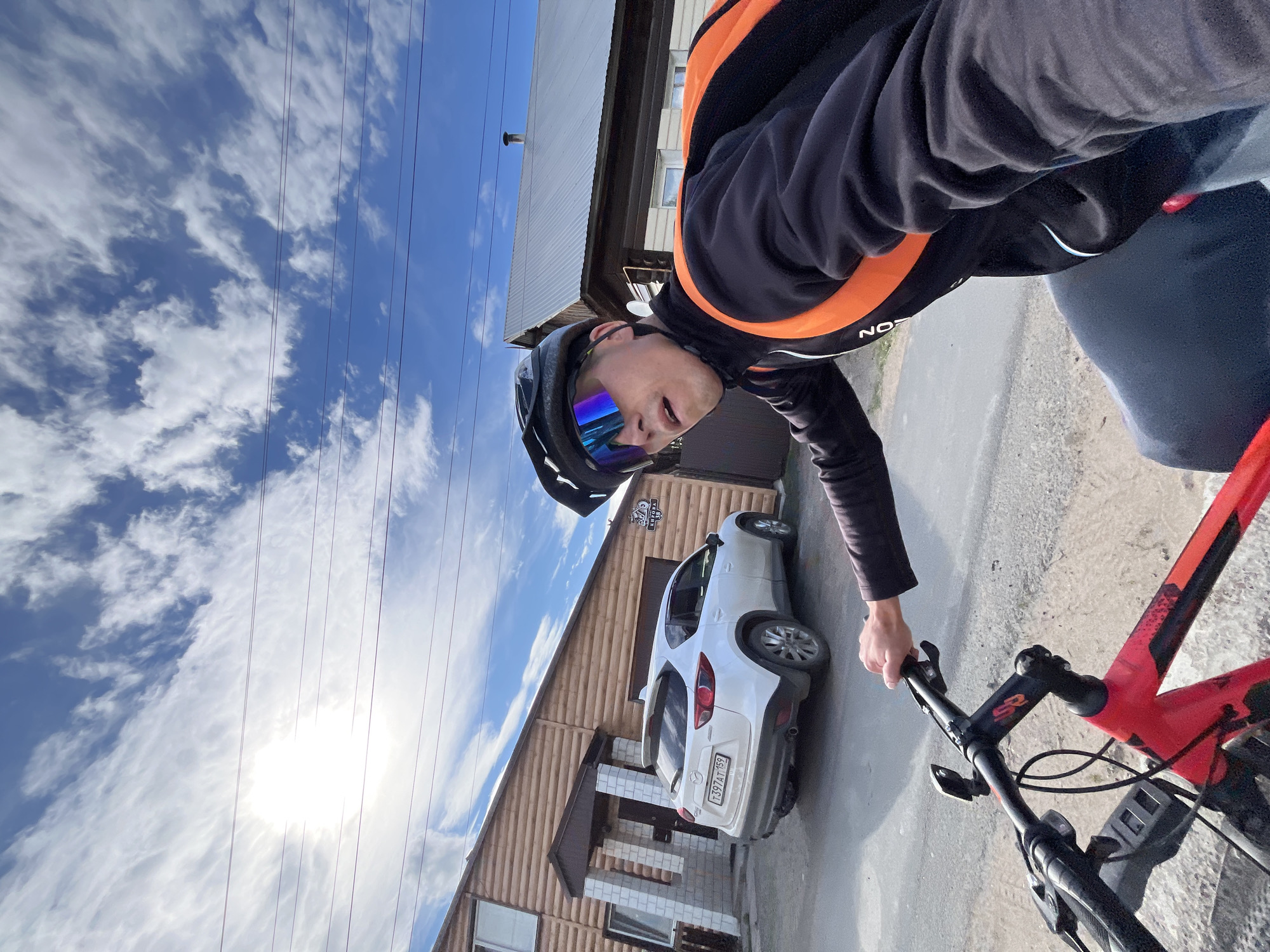 A person is riding a bicycle and filming themselves with a forward-facing camera. The cyclist is wearing a black helmet, reflective sunglasses, and a black jacket with orange accents. They are holding onto the handlebars of a red and black bicycle. The scene appears to be a residential street, with a wooden building and a white car visible in the background. The sky is blue with some wispy clouds, and the sun is shining brightly. The time of day appears to be daytime. The overall mood is active and outdoorsy. Visible text on the car's license plate reads "T397AT 159" and on a street sign reads "ул. Мирная 34".