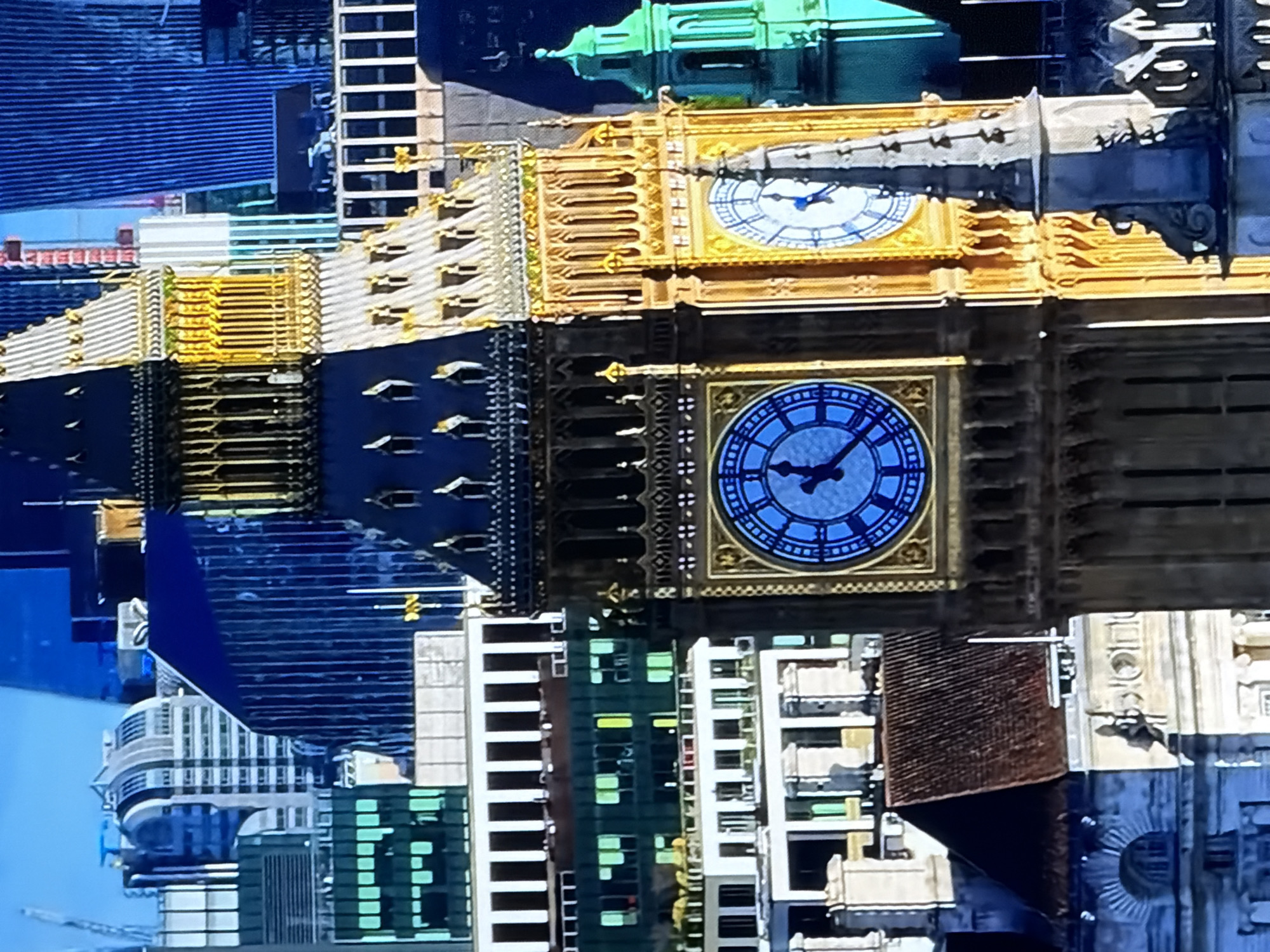 The image depicts the Elizabeth Tower, commonly known as Big Ben, located in London, United Kingdom. The tower's two visible clock faces display different lighting conditions and subtly varying clock face colors. The left clock face features a deep blue background with white Roman numerals, showing time approximately 1:58. The right clock face has a light blue and white background with black Roman numerals, also indicating approximately 1:58. The left portion of the tower is in shadow, presenting darker stonework, while the right portion is brightly illuminated by sunlight, exhibiting a golden hue on its stone and ornate architectural details. In the background, a dense urban cityscape is visible, featuring various multi-story buildings, including modern structures with reflective blue and green glass facades, older white buildings, and one with a distinctive rounded roof. No individuals are discernible in the scene. The sky is clear and blue.