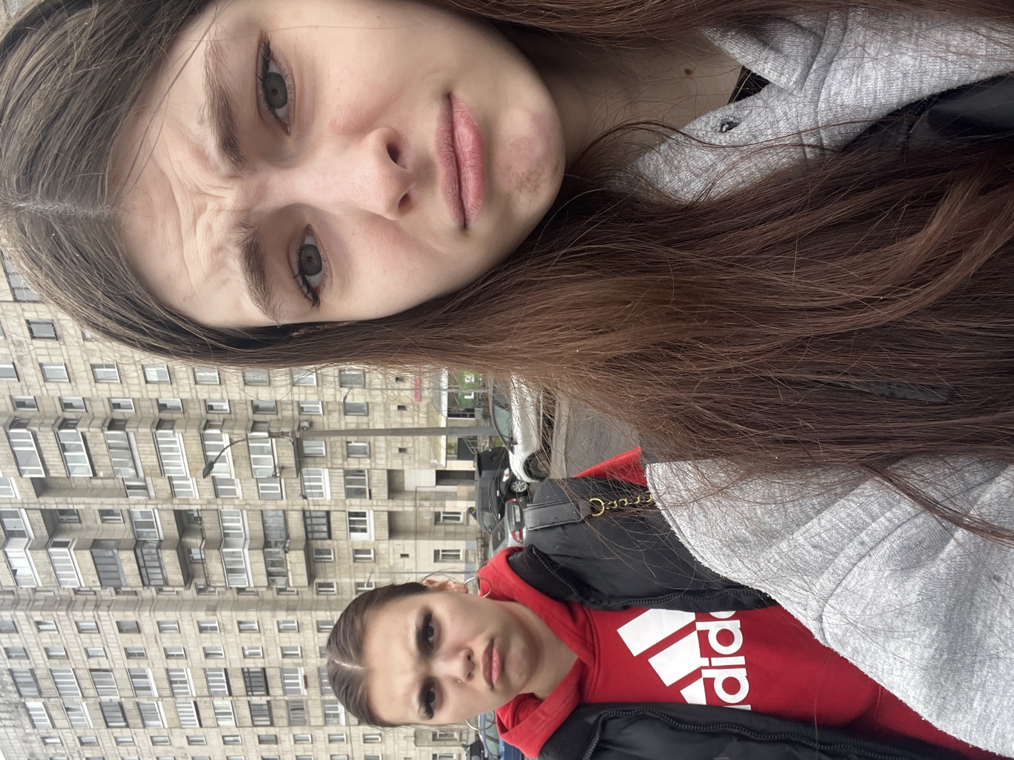 The image shows two women in an urban setting. The woman in the foreground has long brown hair, a grey hoodie, and a black jacket and appears to be taking a selfie. Her face expresses disapproval. The second woman is wearing a red Adidas hoodie and a black jacket. Her hair is in a ponytail. She appears to be making a similar facial expression. The background features a Soviet-era apartment building, a lamppost, and parked cars. The apartment building is in a style typical of structures found in Saint Petersburg, Russia.