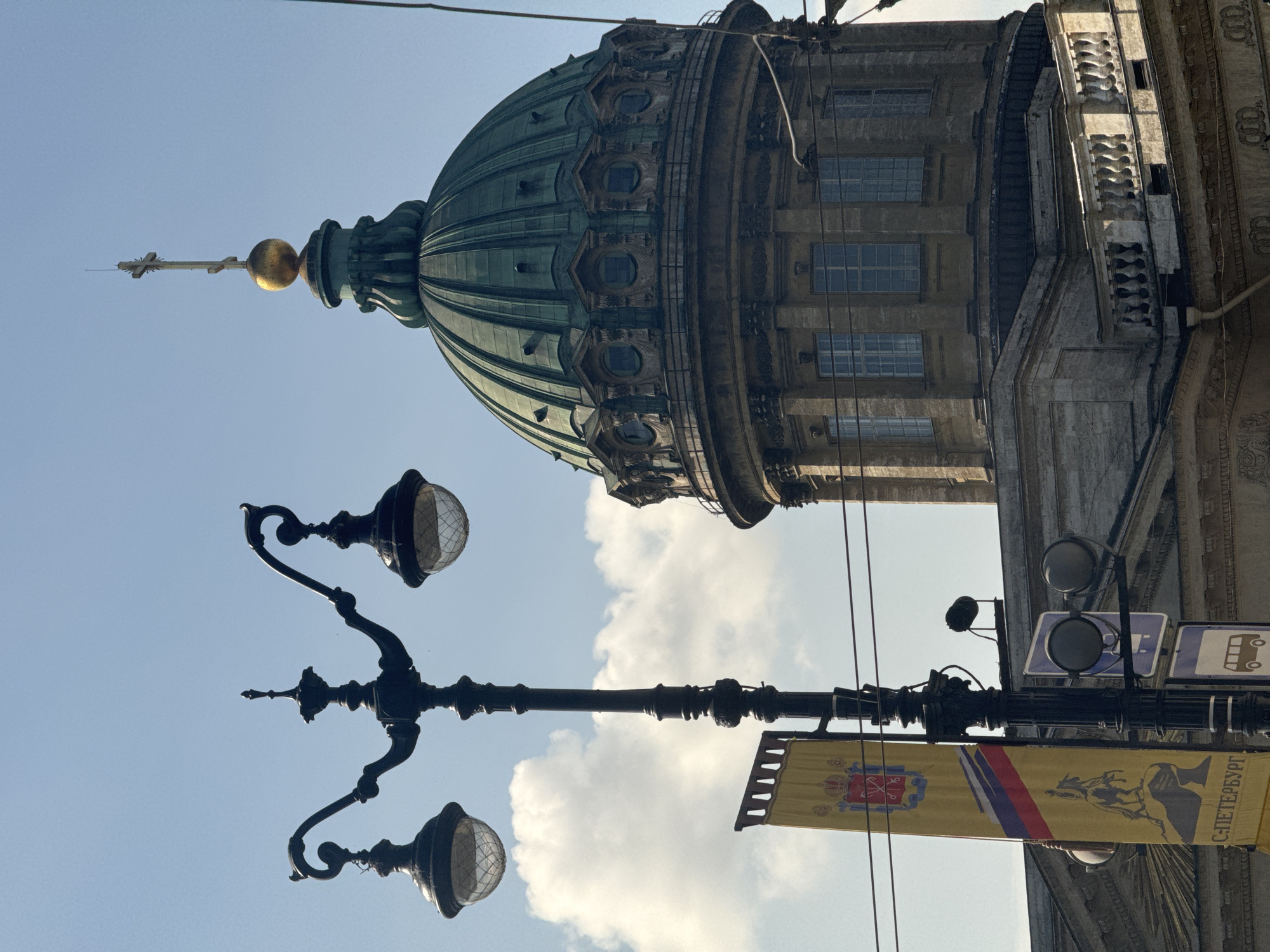 This is a medium-shot, eye-level, outdoor photograph of the Kazan Cathedral in Saint Petersburg, Russia, on a clear day with some clouds. In the foreground, a black wrought-iron lamppost with two glass globe lights is positioned to the left of the frame. To the left of the lamppost is a yellow banner with a red and blue stripe and an image of a person on horseback. The banner also has text at the bottom that reads "С-ПЕТЕРБУРГ". The Kazan Cathedral, a prominent landmark in Saint Petersburg, is visible in the background on the right. Its large, green dome with a gold orb and cross at the apex dominates the upper right portion of the image. The cathedral's stone facade, columns, and windows are also visible. The sky is blue with fluffy white clouds. The image appears to have been taken from a street level, with the lamppost and banner indicating a public urban setting.