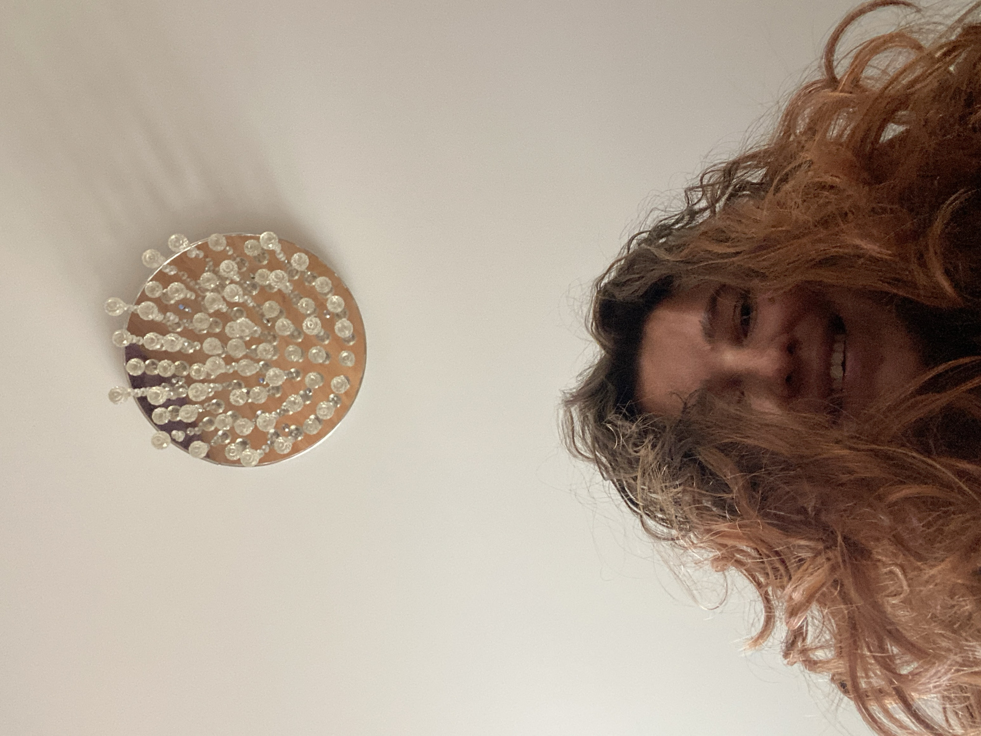 An extreme low-angle, upward-facing outdoor shot captures a person's head and face from below, positioned in the lower half of the frame. Their curly, reddish-brown hair cascades around their face, obscuring much of it. A bright smile is visible, with teeth showing, but the eyes are not clearly discernible. Above the person's head, a circular light fixture with numerous clear, bulb-like ornaments is mounted on a white ceiling, which is consistent with an indoor setting, potentially within a residential apartment or a commercial establishment in the city of Perm, Russia. The lighting is somewhat dim, casting faint shadows from the fixture onto the ceiling. The overall impression is a candid, informal snapshot taken from an unusual perspective.