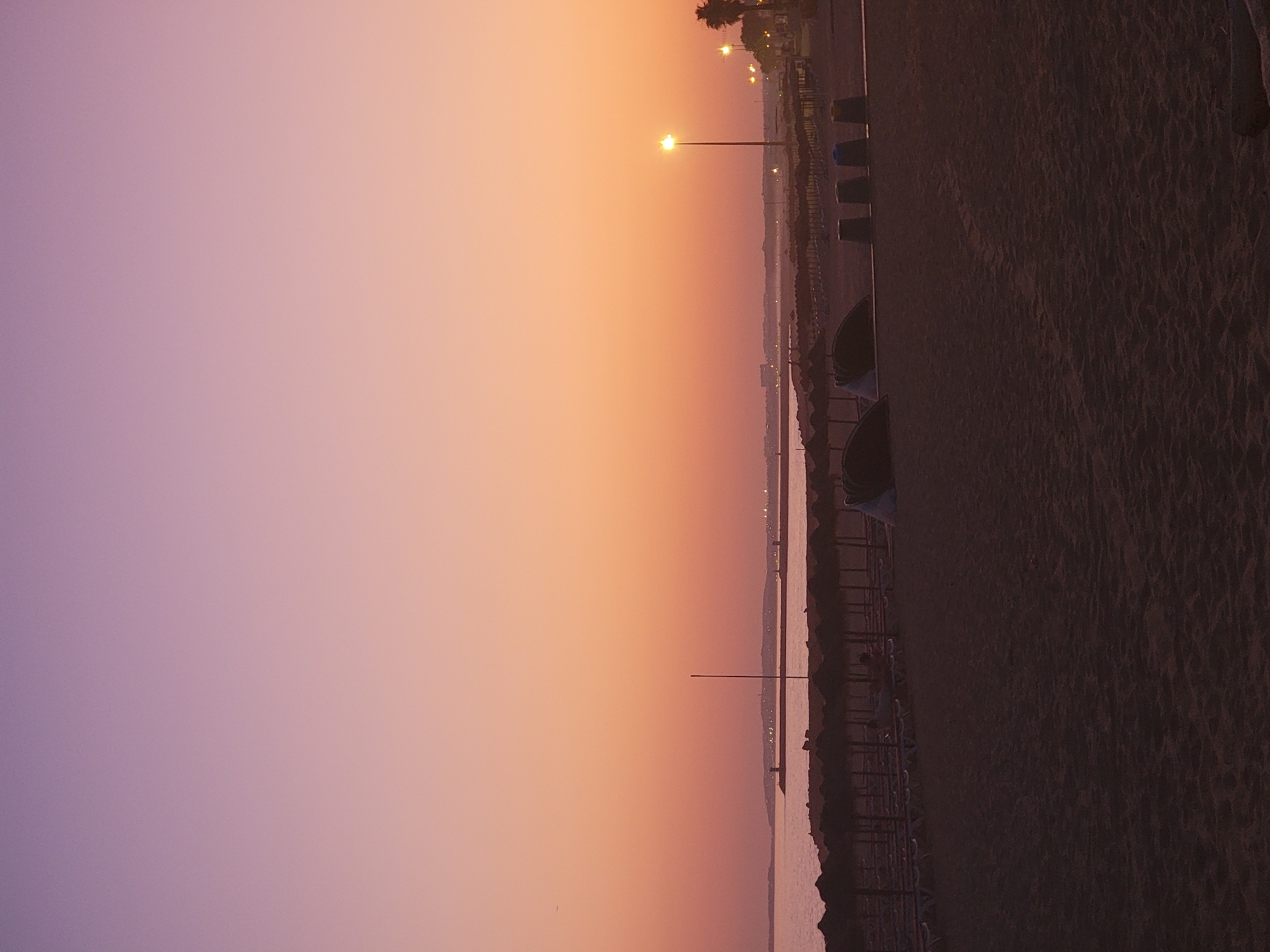 The image depicts a beach scene at dusk in Quarteira, Portugal. The sky exhibits a gradient of soft purple to pink and orange hues, indicative of a sunset. In the foreground, the sandy beach is visible with tire tracks left by vehicles or machinery. A row of thatched umbrellas and lounge chairs stretches across the middle ground, most of which are empty and appear set up for the day's end. Several large dark cylindrical objects, possibly trash receptacles, are lined up near the umbrellas. In the background, the sea is calm, reflecting the colors of the sky. Along the distant horizon, a landmass with buildings and lights is visible, possibly the Quarteira coastline. A single lamppost with its light illuminated stands on the right side of the image.