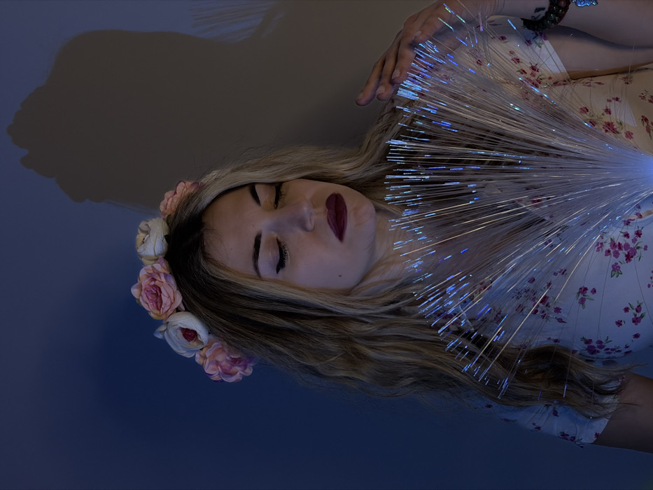 An indoor portrait taken in Budva, Montenegro, features an adult female positioned against a plain, dimly lit wall. The subject has long, wavy blonde hair and wears a floral crown consisting of pink and white artificial roses. Her eyes are closed, and she has dark red lipstick applied. She is dressed in a light-colored garment with a pink floral pattern. Her left hand is resting on an illuminated optical fiber fan, which emits blue and white light, casting a subtle glow onto her face and upper body. A beaded bracelet with a charm is visible on her left wrist. A faint shadow, consistent with a human profile, is cast on the upper right portion of the background wall.